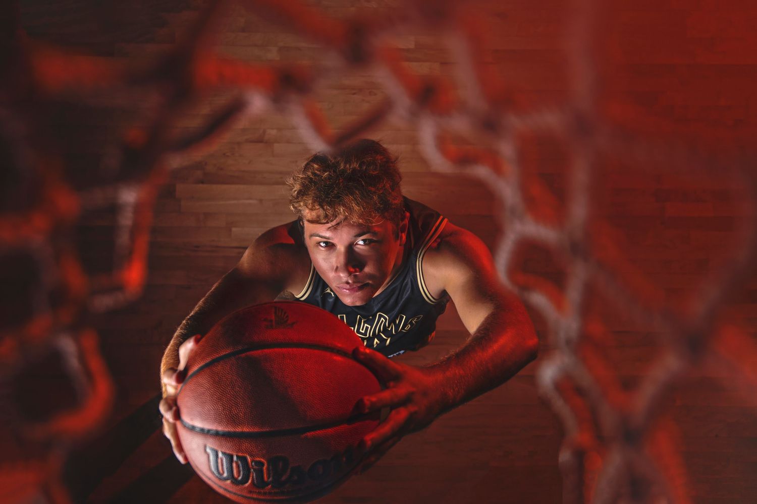 Basketball player holding Wilson ball photographed from below through net with red dramatic lighting.