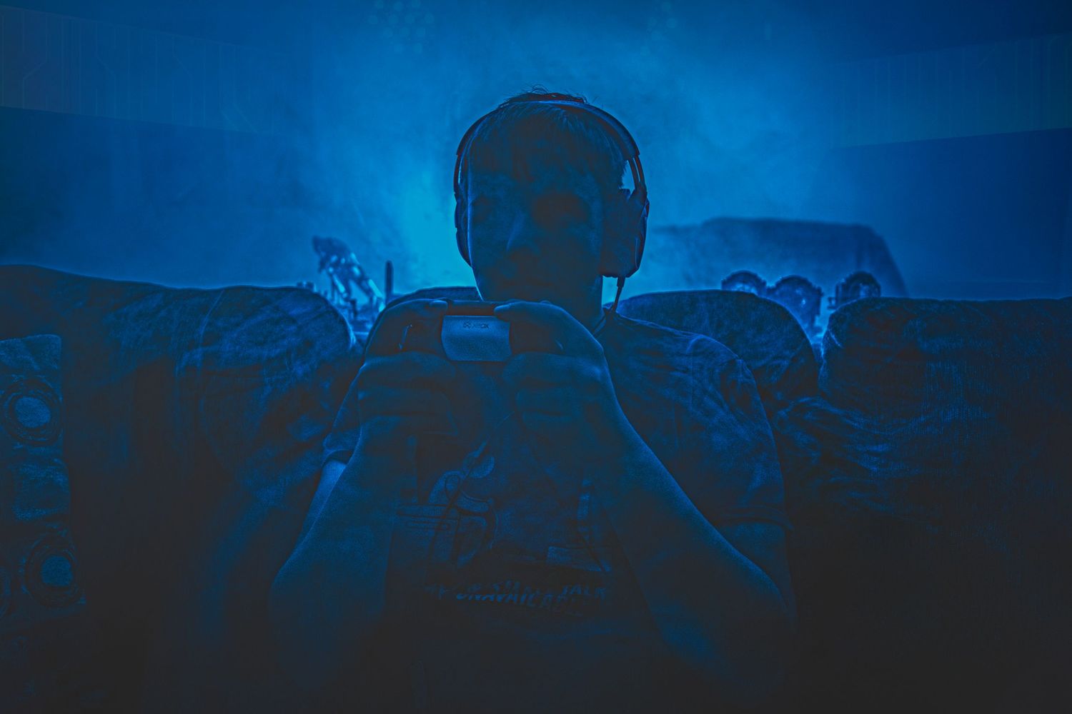 A soldier in tactical gear and headphones sits in a dark blue-toned military vehicle with fellow troops.