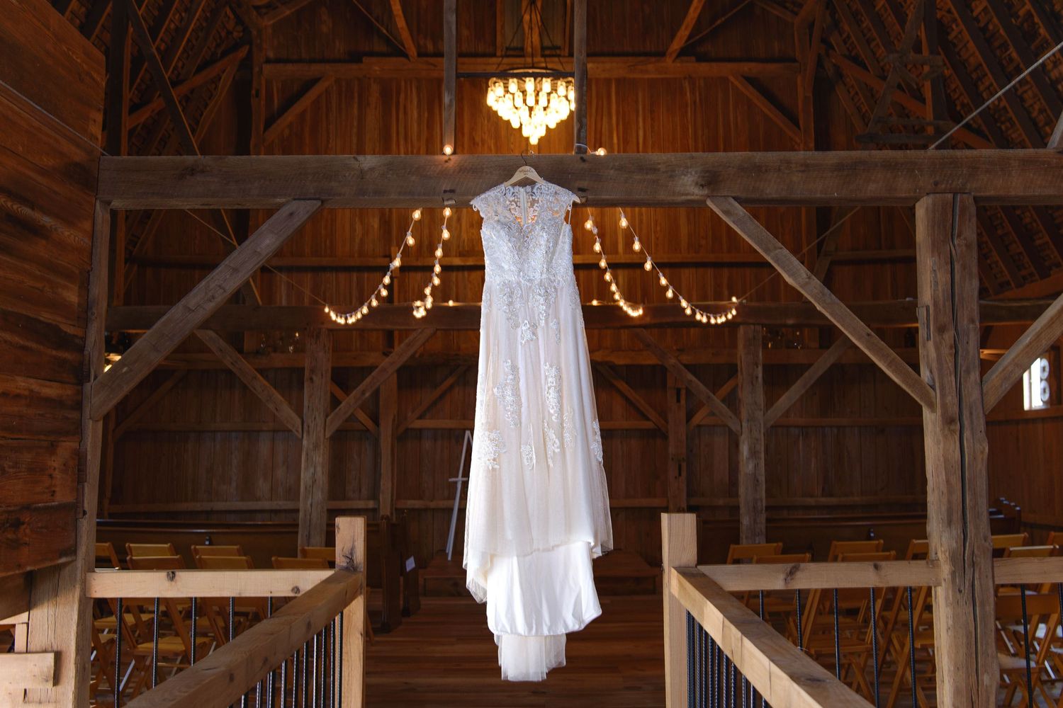 Elegant white lace wedding dress hanging from rustic barn rafters with warm string lights glowing.