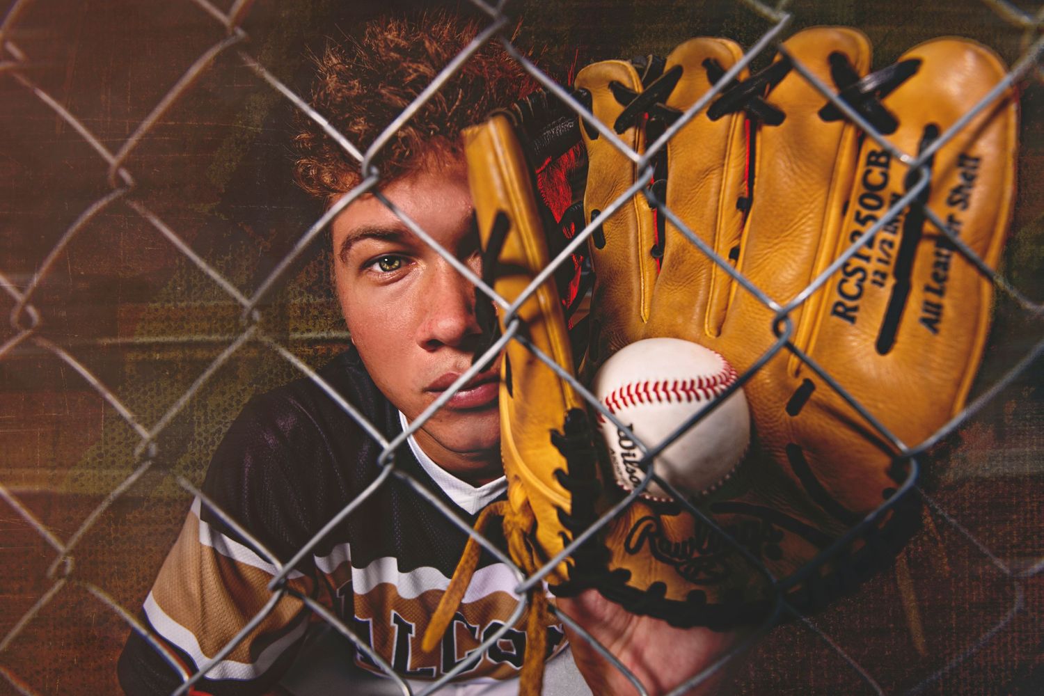 Baseball player holding glove and ball behind chain-link fence in dramatic sports portrait.
