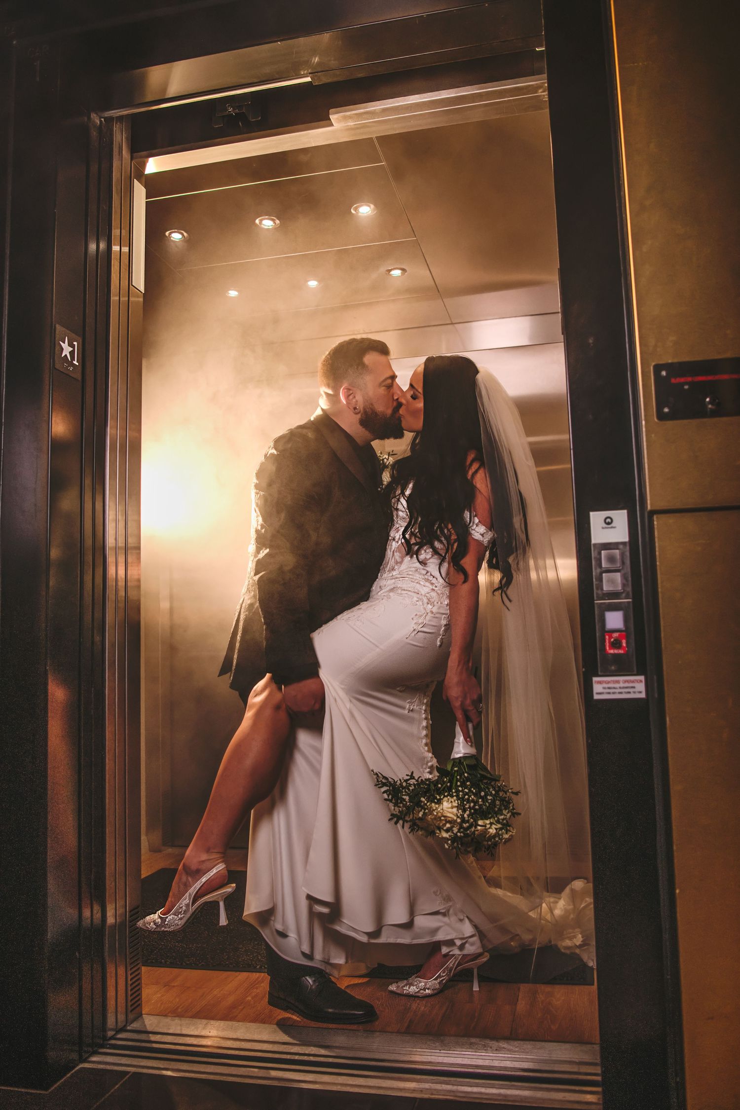 Bride and groom sharing a romantic kiss inside a golden-lit elevator on their wedding day.