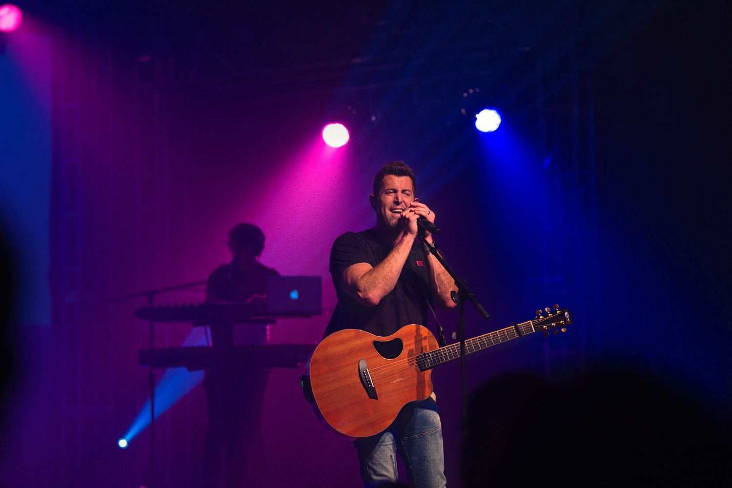 Male singer performing on stage with guitar, colorful purple and pink stage lights illuminating the concert venue.