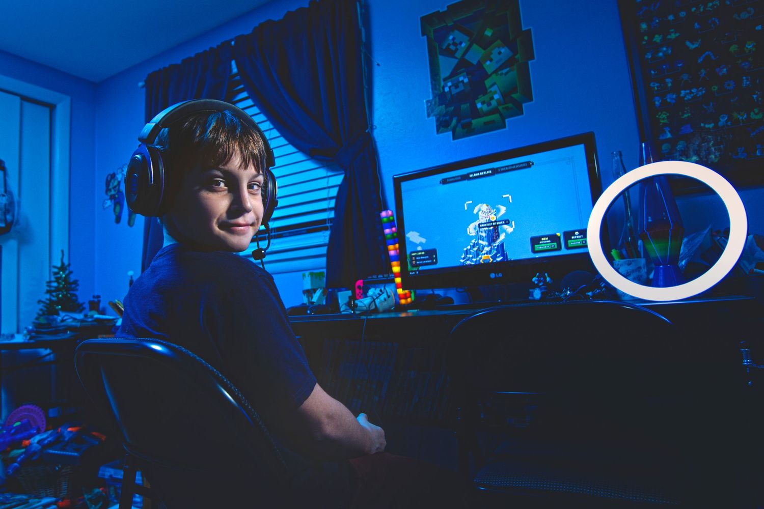 A smiling boy wearing headphones sits at a gaming setup with a glowing ring light in a blue-lit room.