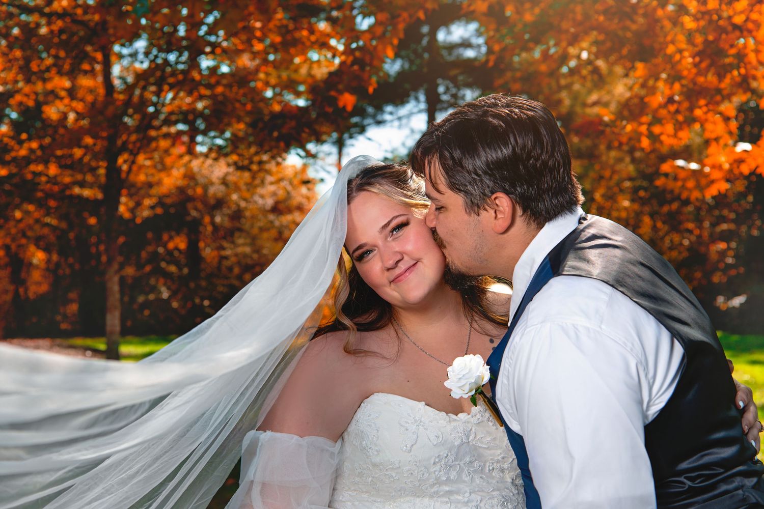 Groom kissing bride on the cheek outdoors surrounded by vibrant orange and red autumn foliage.
