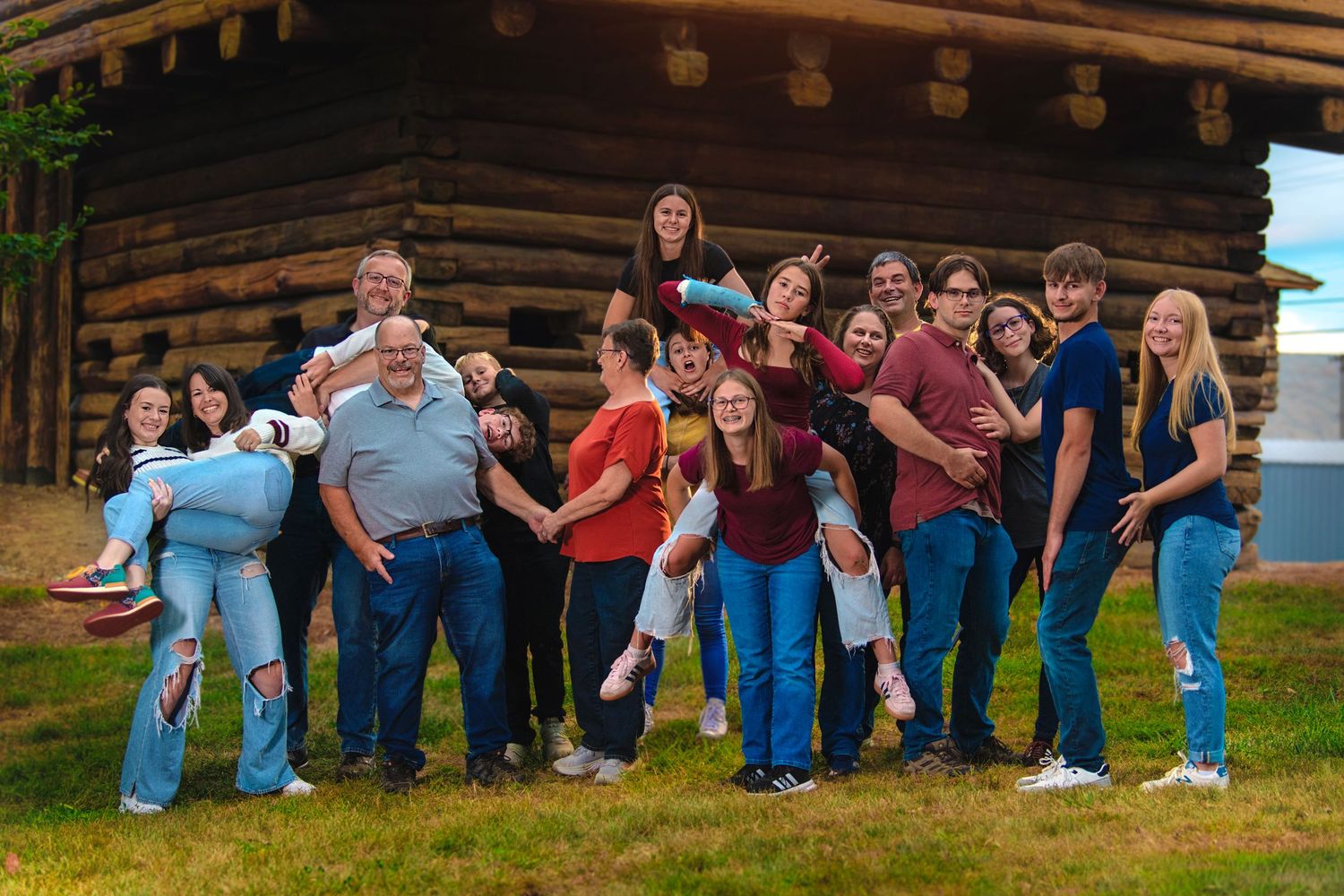 Large family group posing playfully outside a rustic log cabin on green grass during a fall outdoor photo session.