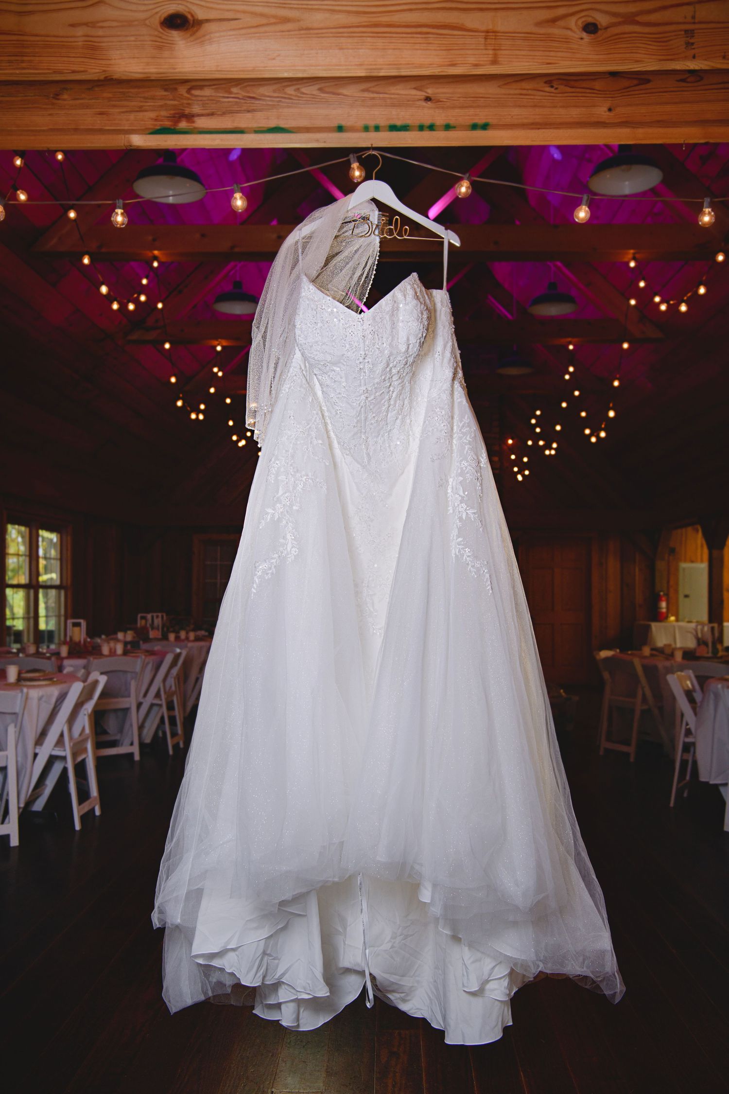 Elegant white lace wedding dress with veil hanging in a rustic barn reception venue with string lights.