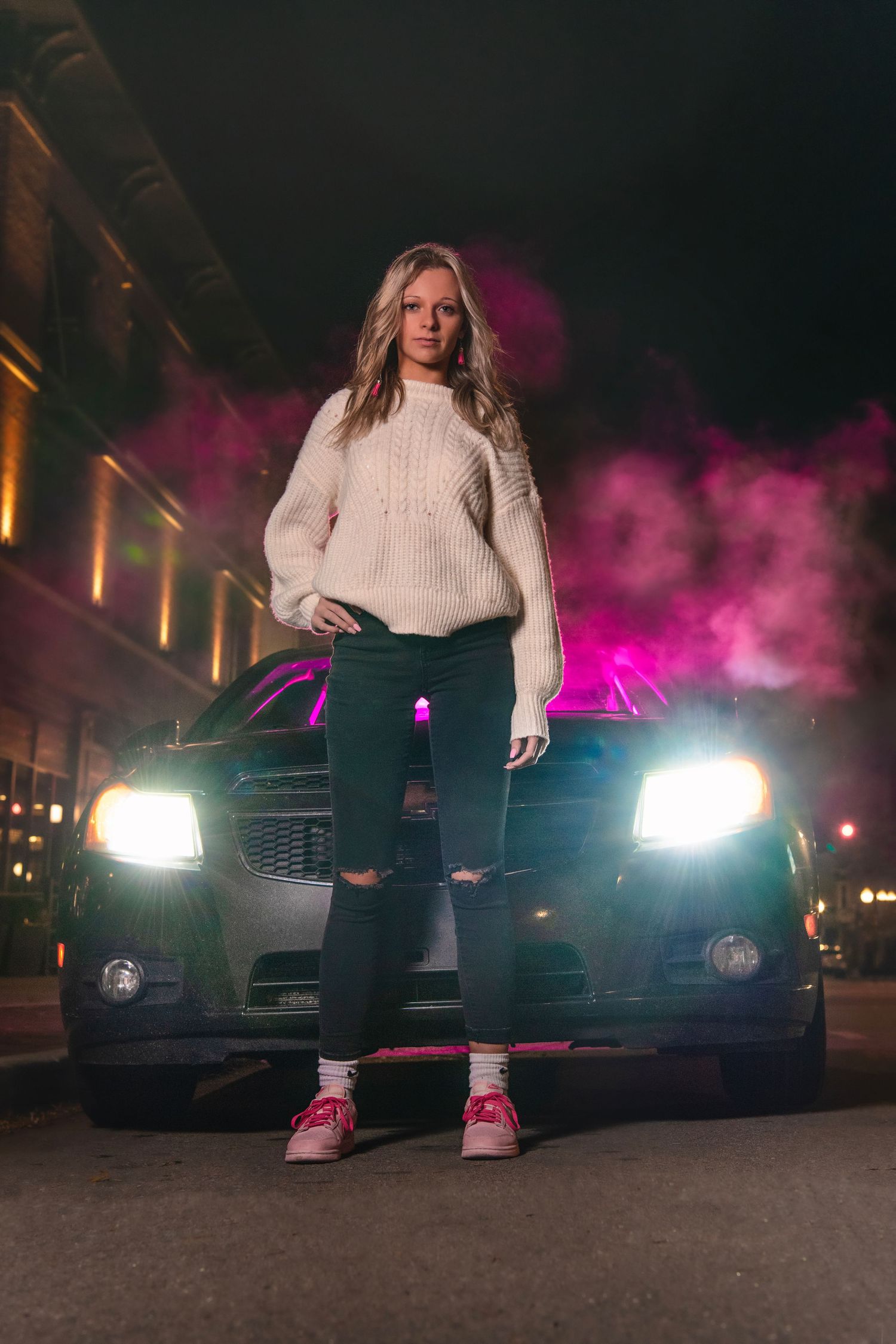 Blonde woman in white sweater standing in front of car headlights with pink smoke at night.