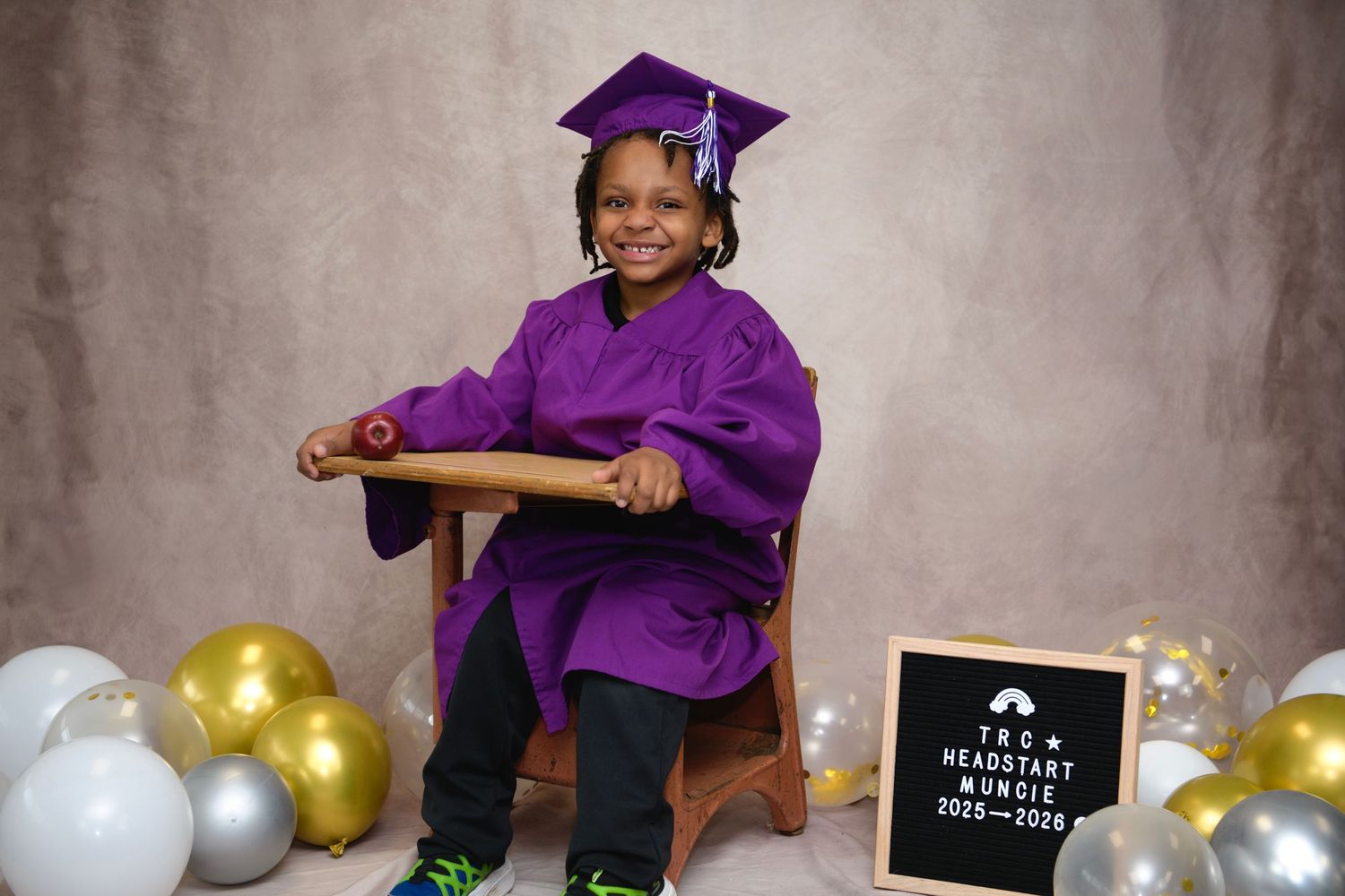 Young child in purple graduation cap and gown seated with balloons and a TRC Head Start 2025 sign.
