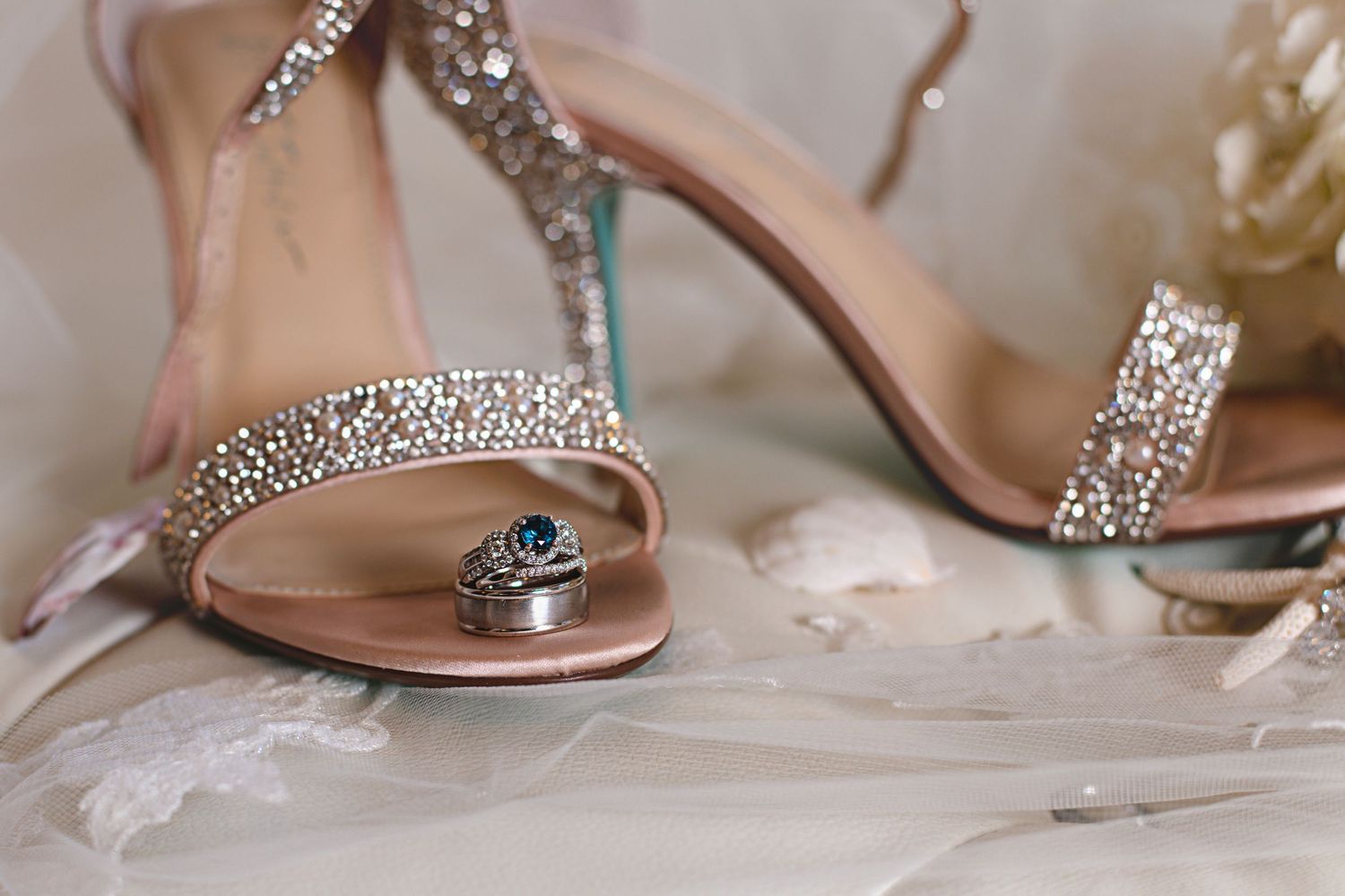 A silver ring with blue gemstone resting against crystal-embellished nude bridal heels on white lace.