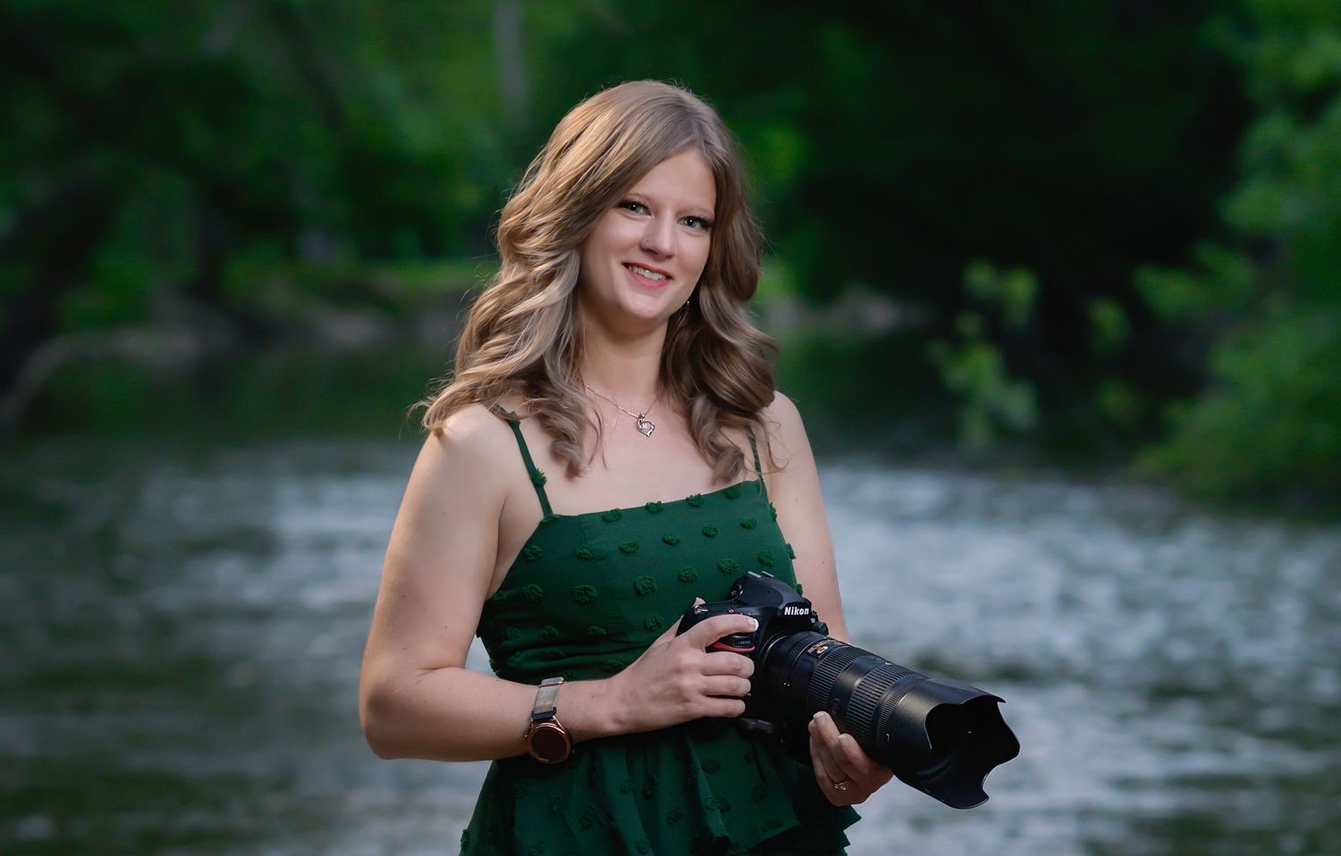 Smiling blonde woman in green dress holding a professional camera outdoors near a serene river backdrop.