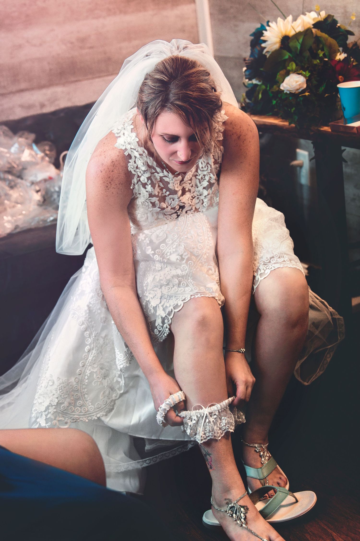 Bride in short lace wedding dress putting on a garter while seated, wearing a veil and sandals.