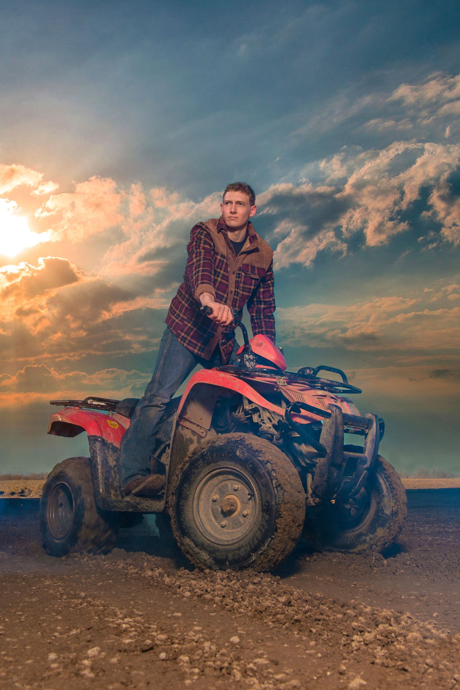 Man in plaid shirt sitting on red ATV quad bike under dramatic sunset sky.