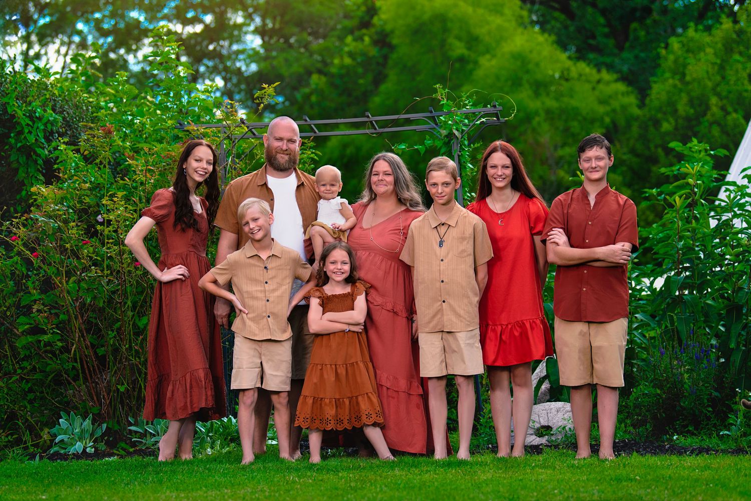 Large family posing outdoors in coordinated earth-tone outfits on a green lawn with lush garden background.