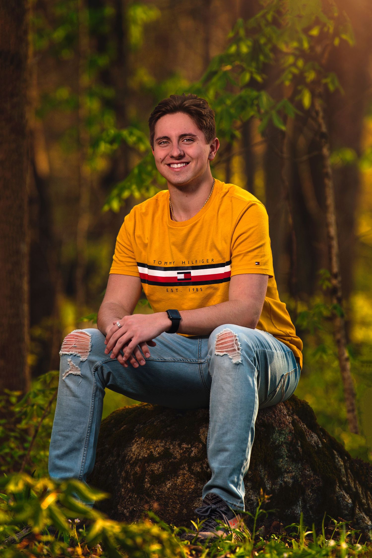 Smiling young man in yellow Tommy Hilfiger shirt sits on rock in sunlit woodland setting.