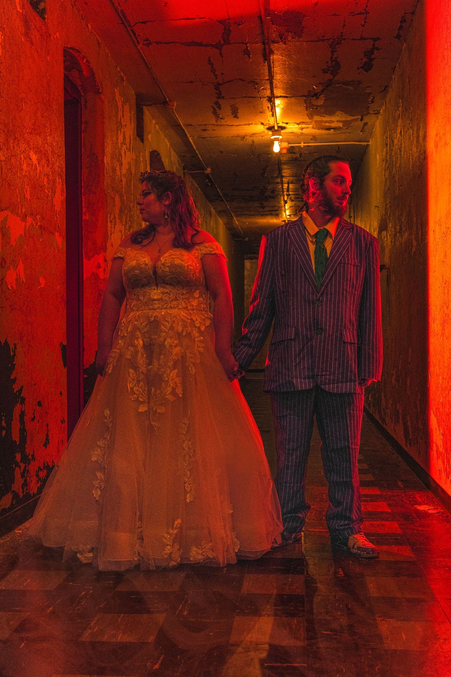 Wedding couple holding hands in a dramatic red-lit hallway, bride in lace gown and groom in pinstripe suit.