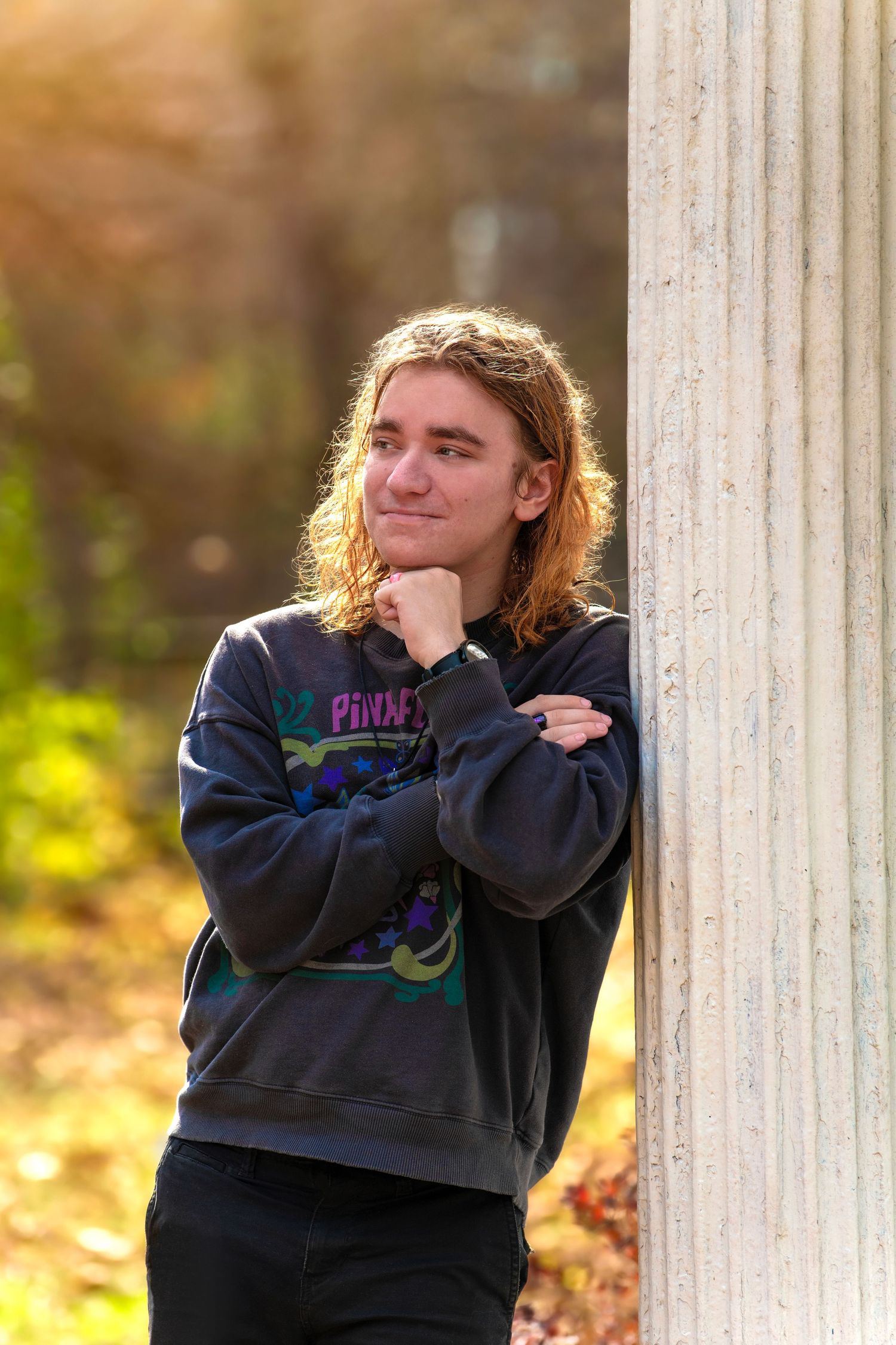 Teen boy with long hair leaning against stone column in sunny autumn outdoor portrait.
