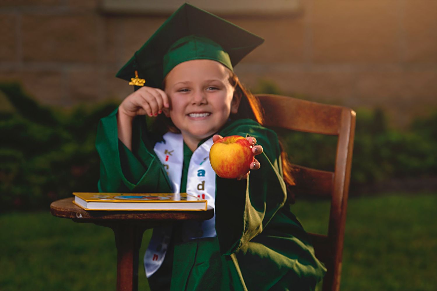 Smiling girl in green graduation cap and gown holds apple while seated at school desk outdoors.