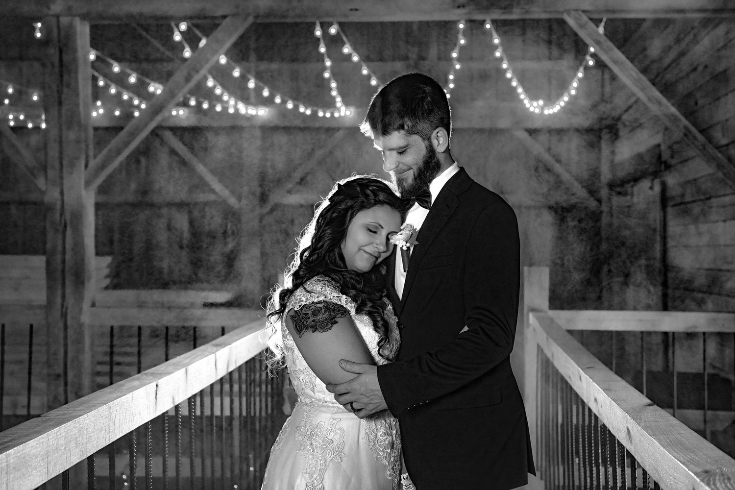 Black and white photo of bride and groom embracing tenderly on a bridge with string lights behind them.