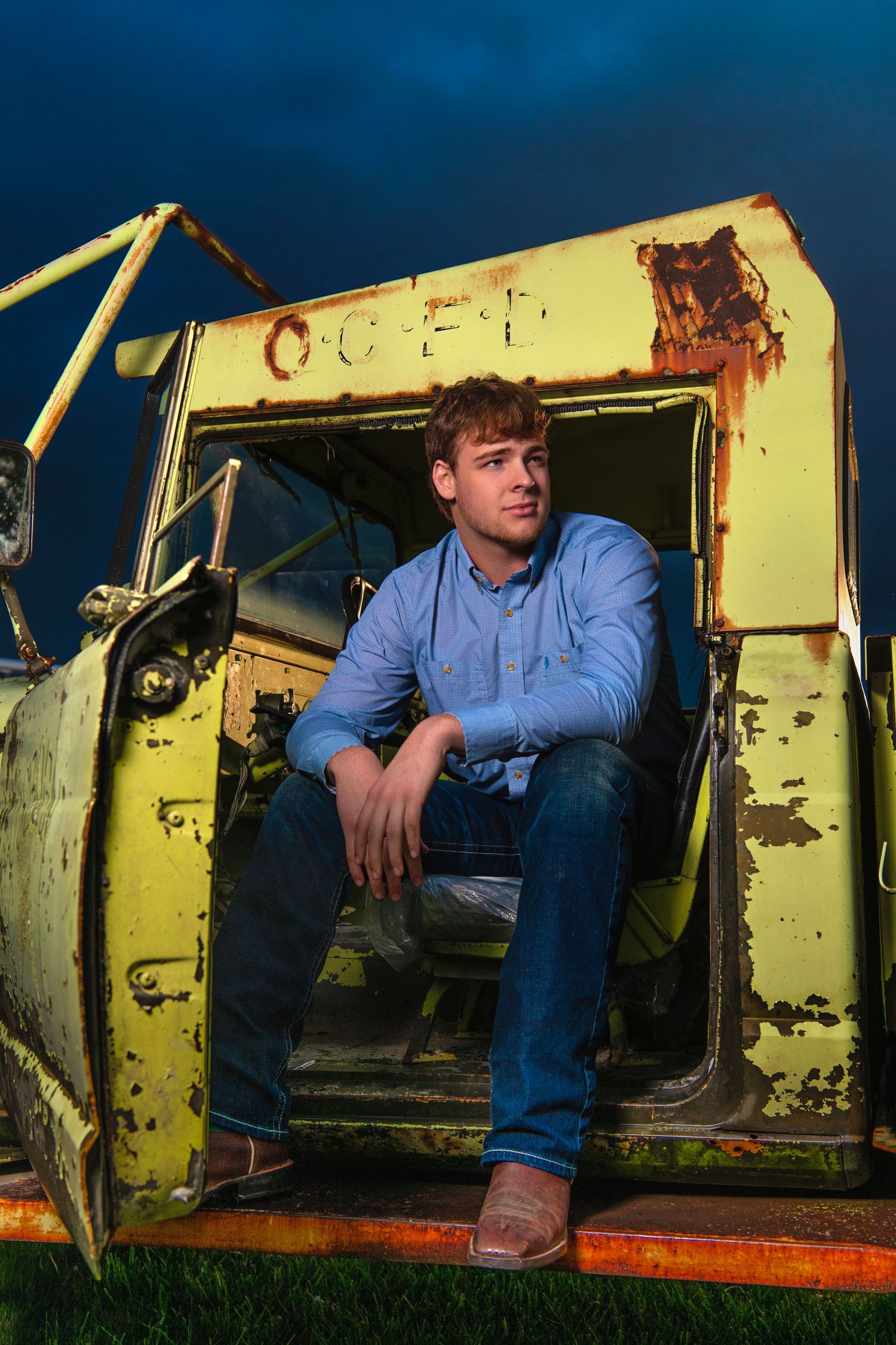 Young man in blue shirt and jeans sitting in rusty abandoned yellow truck under stormy blue sky.