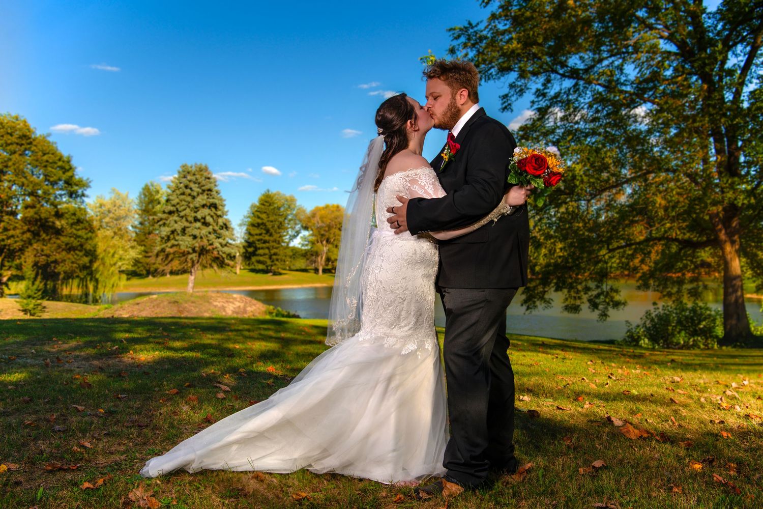 Bride and groom share a romantic kiss outdoors on a sunny autumn day, with colorful fall foliage and a pond in the background.