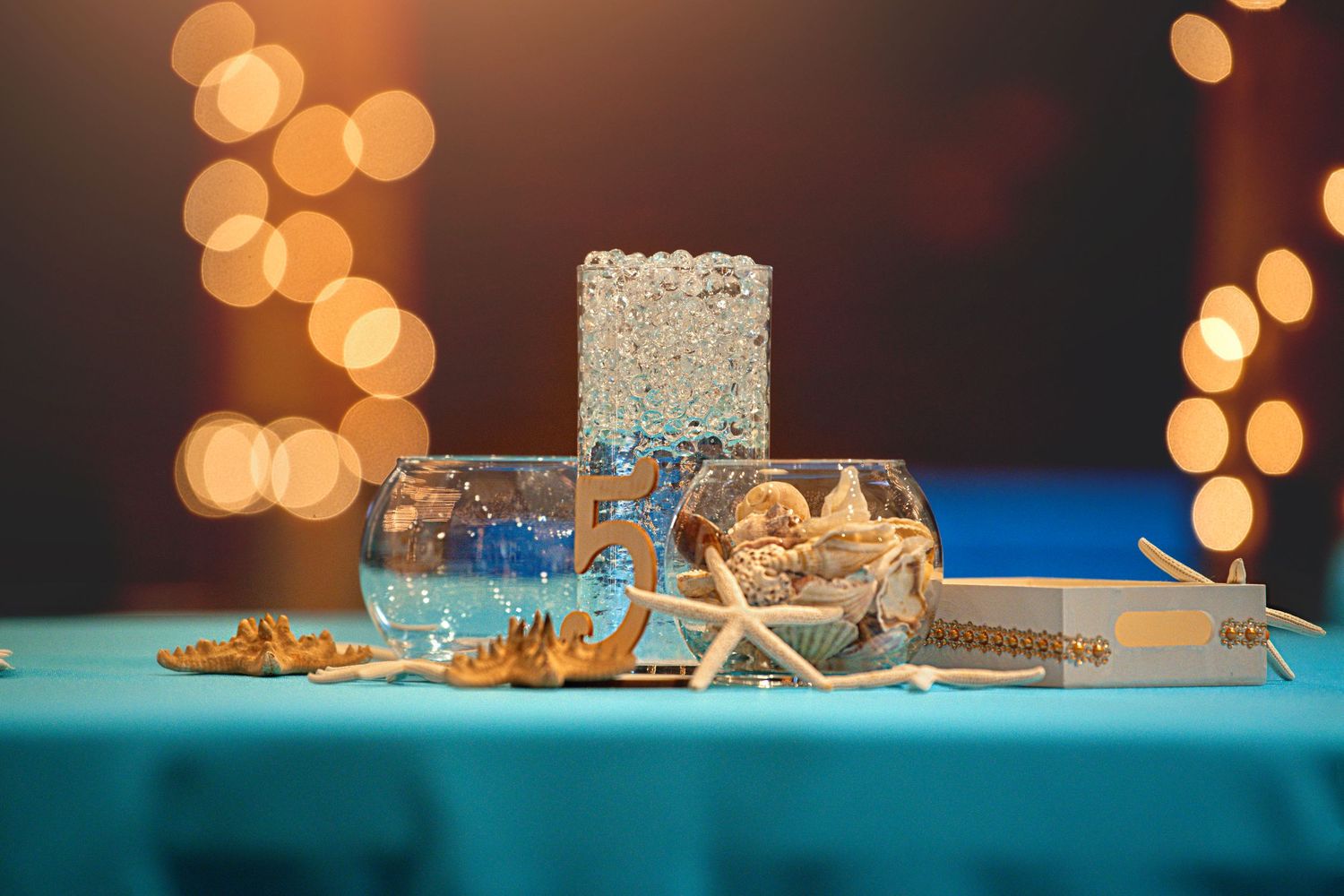 Beach-themed wedding centerpiece with starfish, coral, and glass vases on a teal tablecloth with bokeh lights.