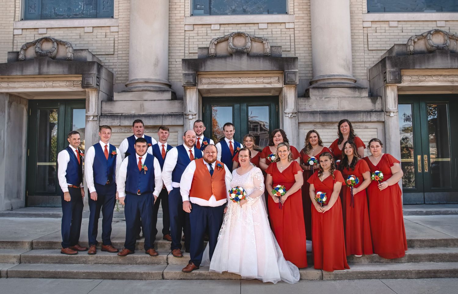 Large wedding party posing on steps of a stone building, bridesmaids in red dresses and groomsmen in navy suits.