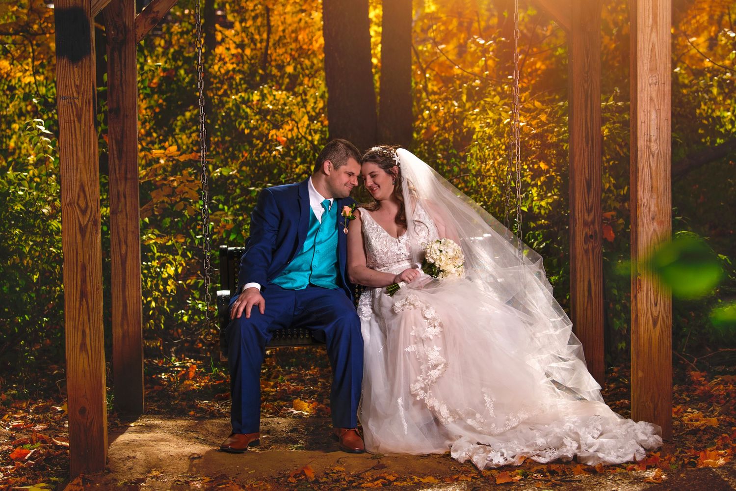 Bride and groom sitting together on a bench surrounded by vibrant autumn foliage in a golden fall forest.