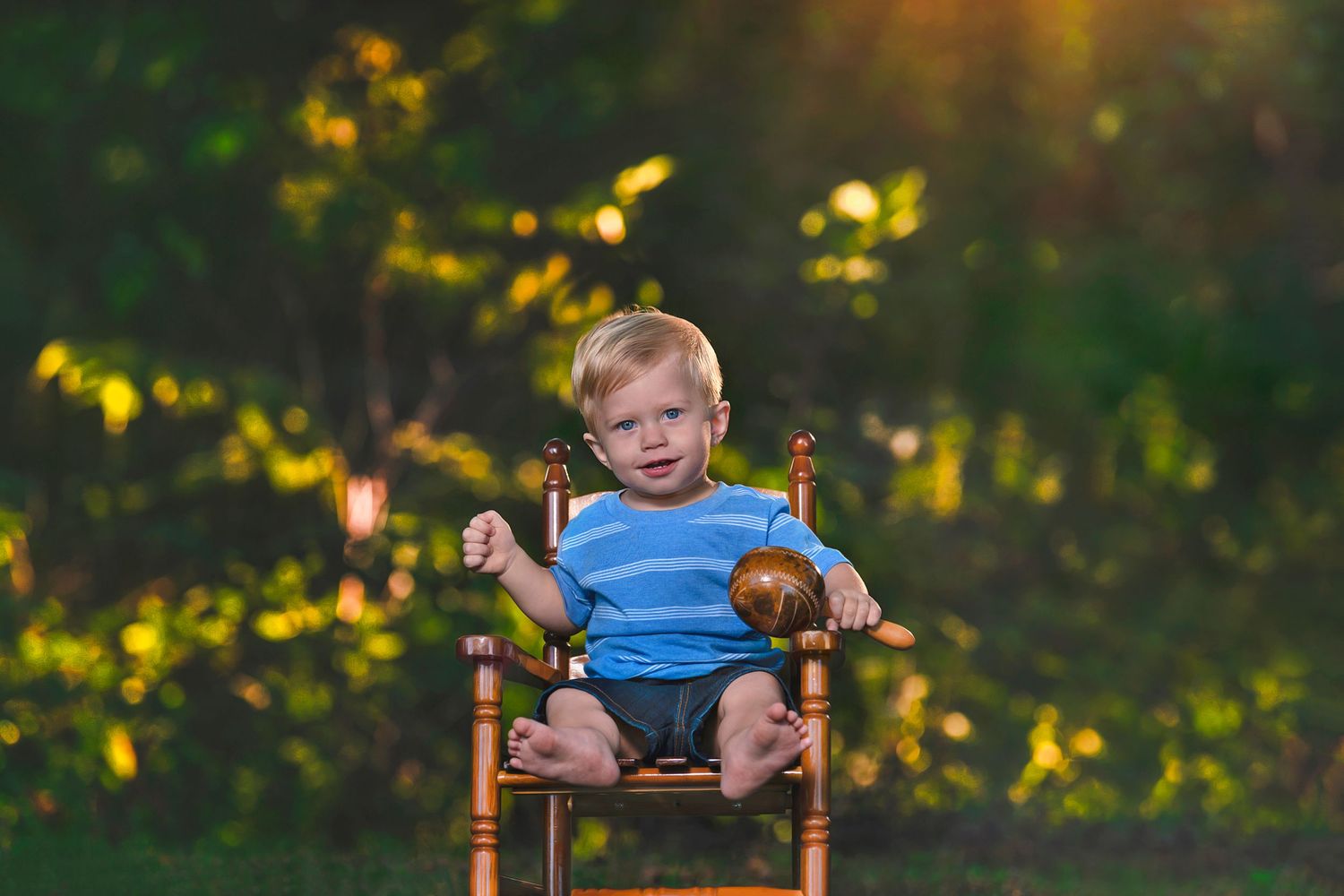 Cute blonde toddler boy sitting in wooden chair holding small pumpkin in sunny green garden.