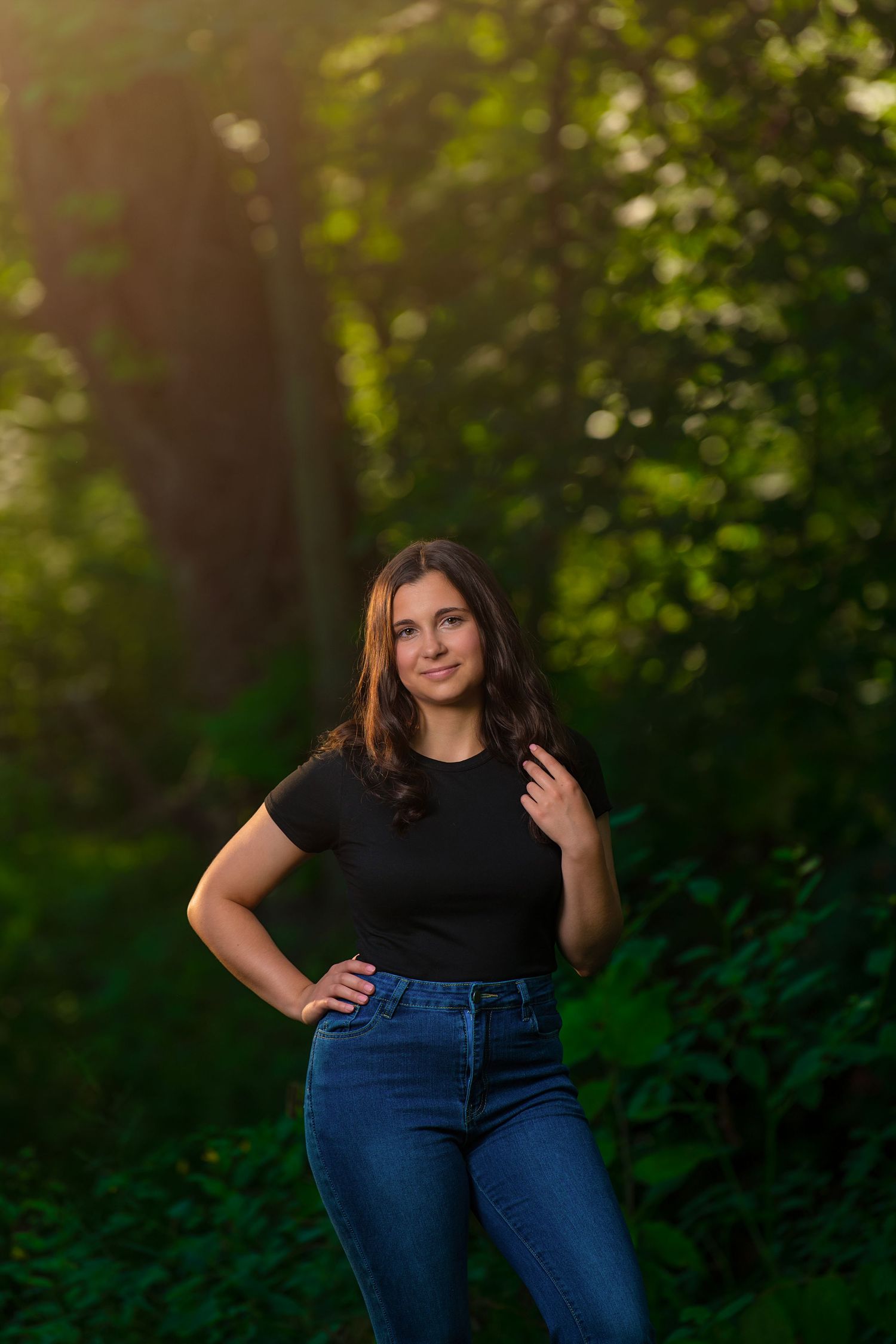 Young woman in black top and jeans poses confidently in sunlit green forest during golden hour.