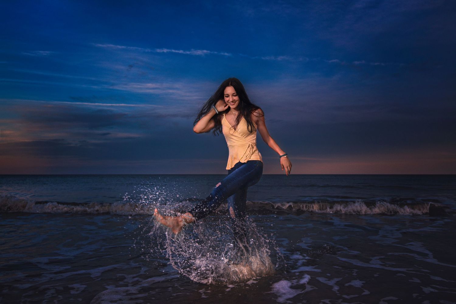 Woman joyfully splashing in ocean waves at sunset, wearing jeans and yellow top on a beach.