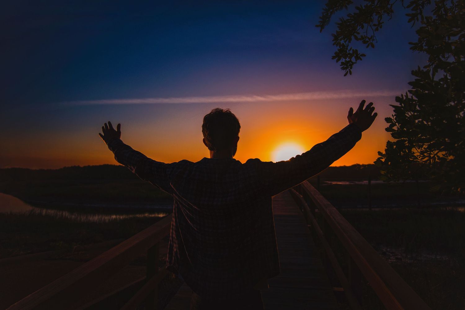 Silhouette of person with arms raised wide open against vibrant orange and blue sunset sky outdoors.