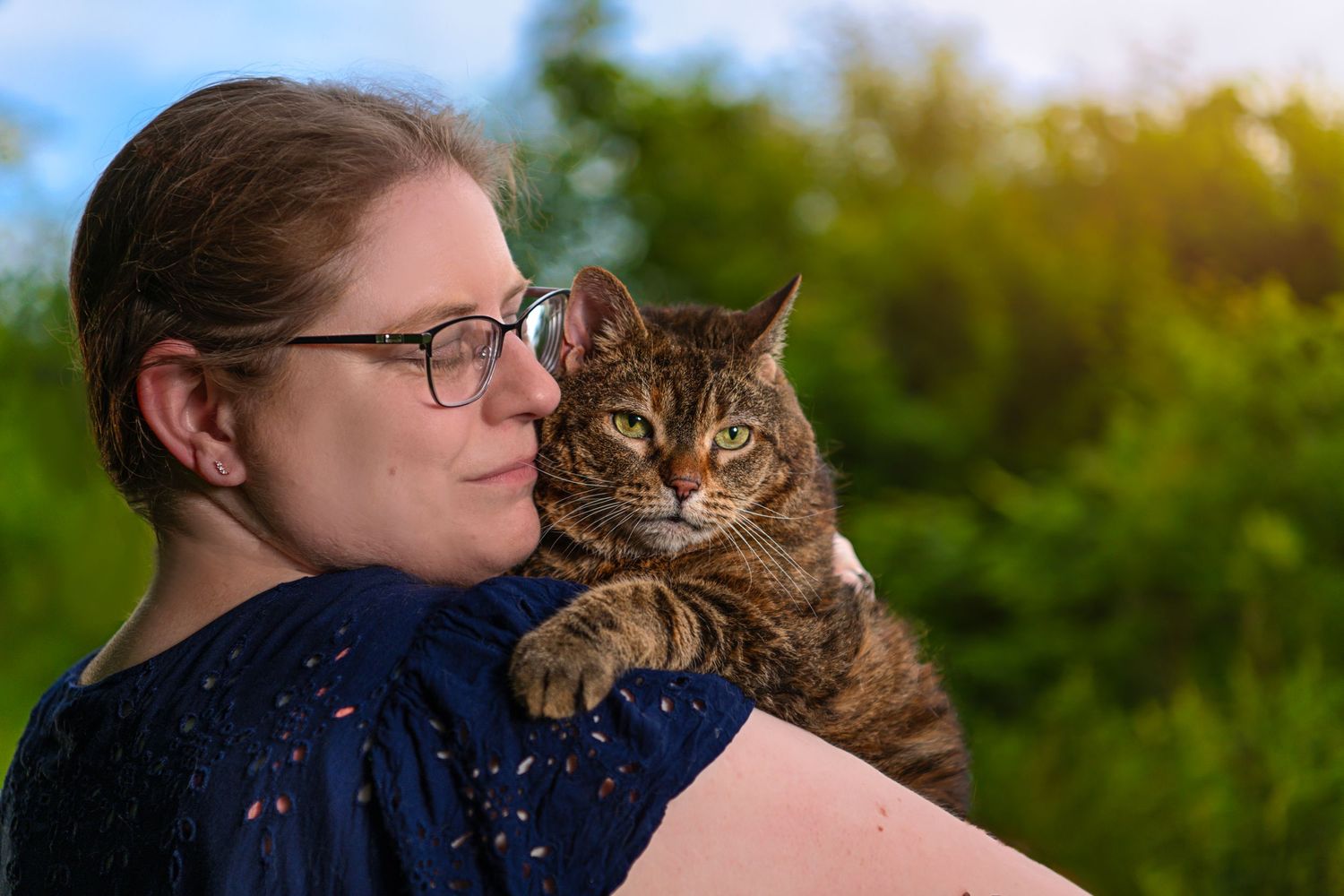 Woman wearing glasses holding a tabby cat outdoors with green foliage background.
