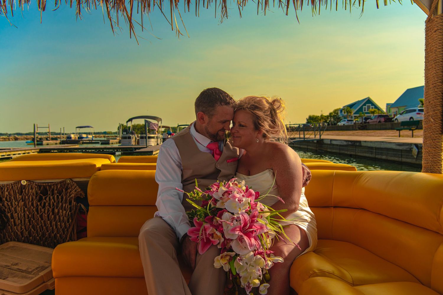 Bride and groom sharing a romantic sunset moment on a yellow boat, bride holding pink tropical bouquet.