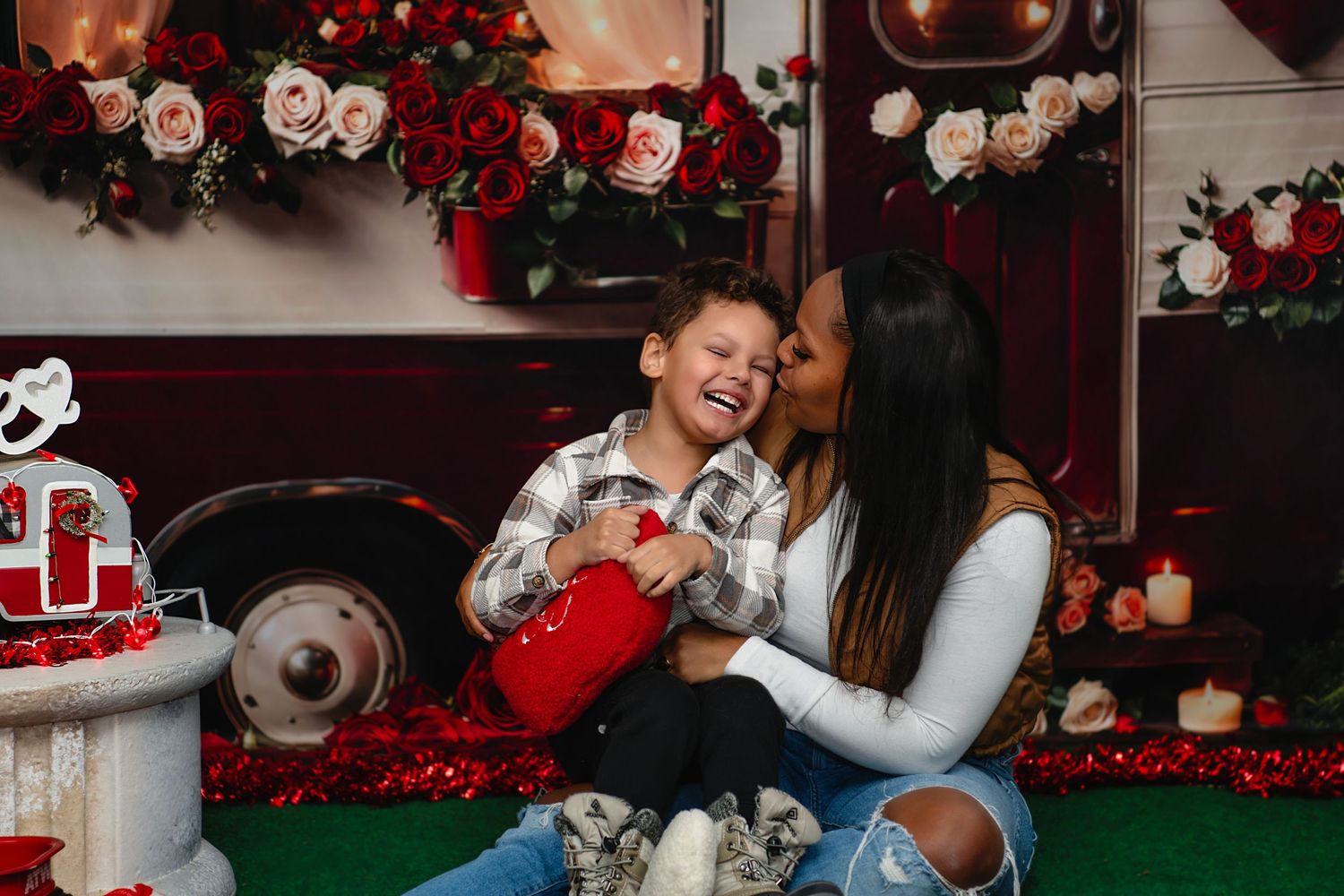 A woman kisses a laughing boy on the cheek during a Valentine's Day photo session with red roses and floral backdrop.