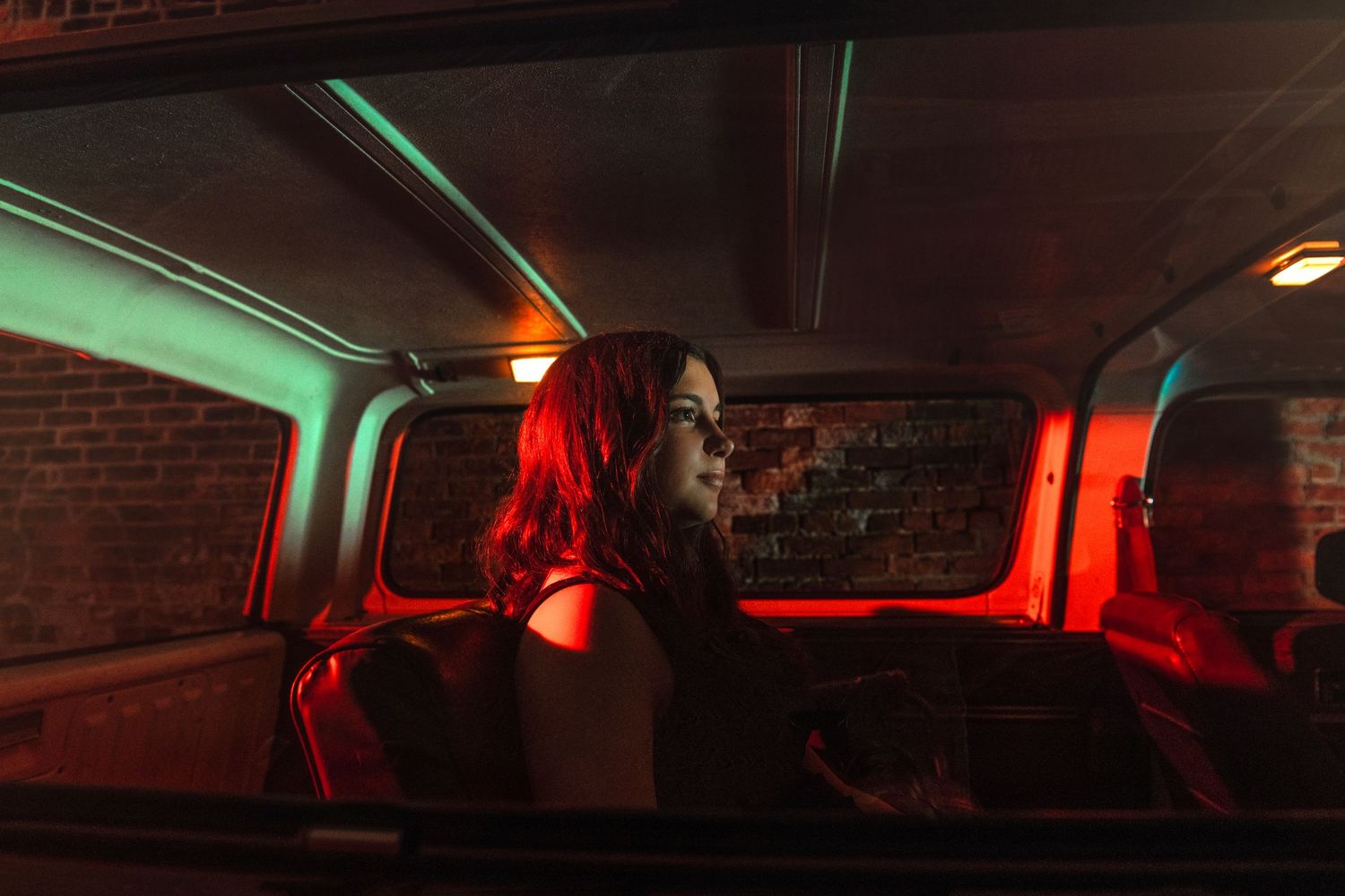 Woman with red hair sitting inside a vintage van illuminated by red and green neon lights at night.