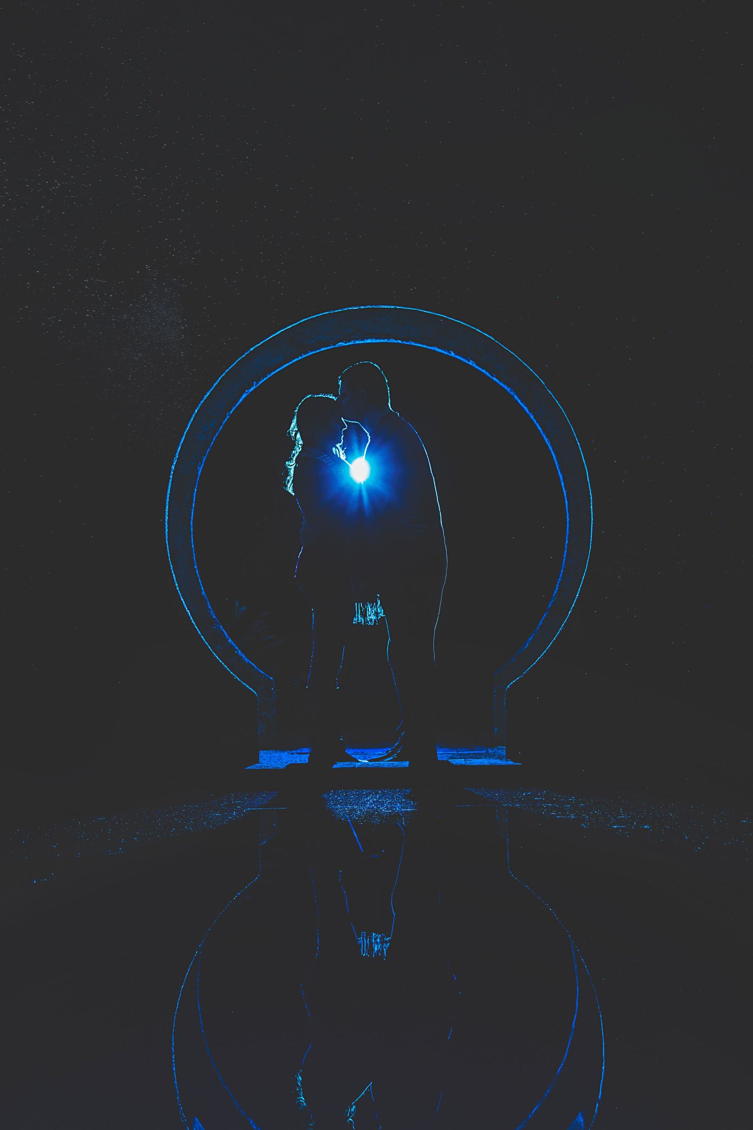 Silhouette of couple kissing inside a blue light-painted circle reflected on wet ground.