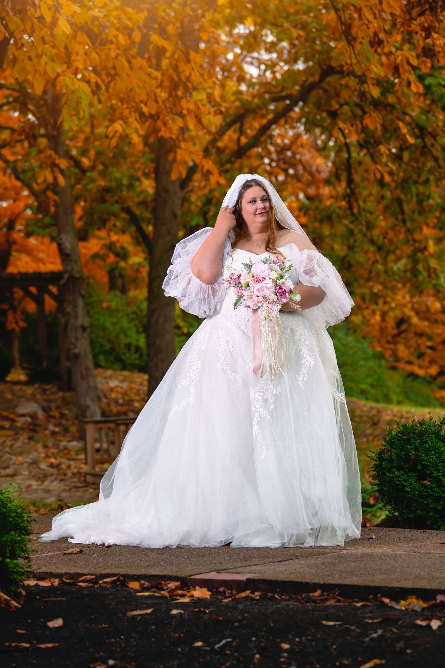 Plus-size bride in a full white ballgown with veil holding a pink bouquet among autumn foliage.