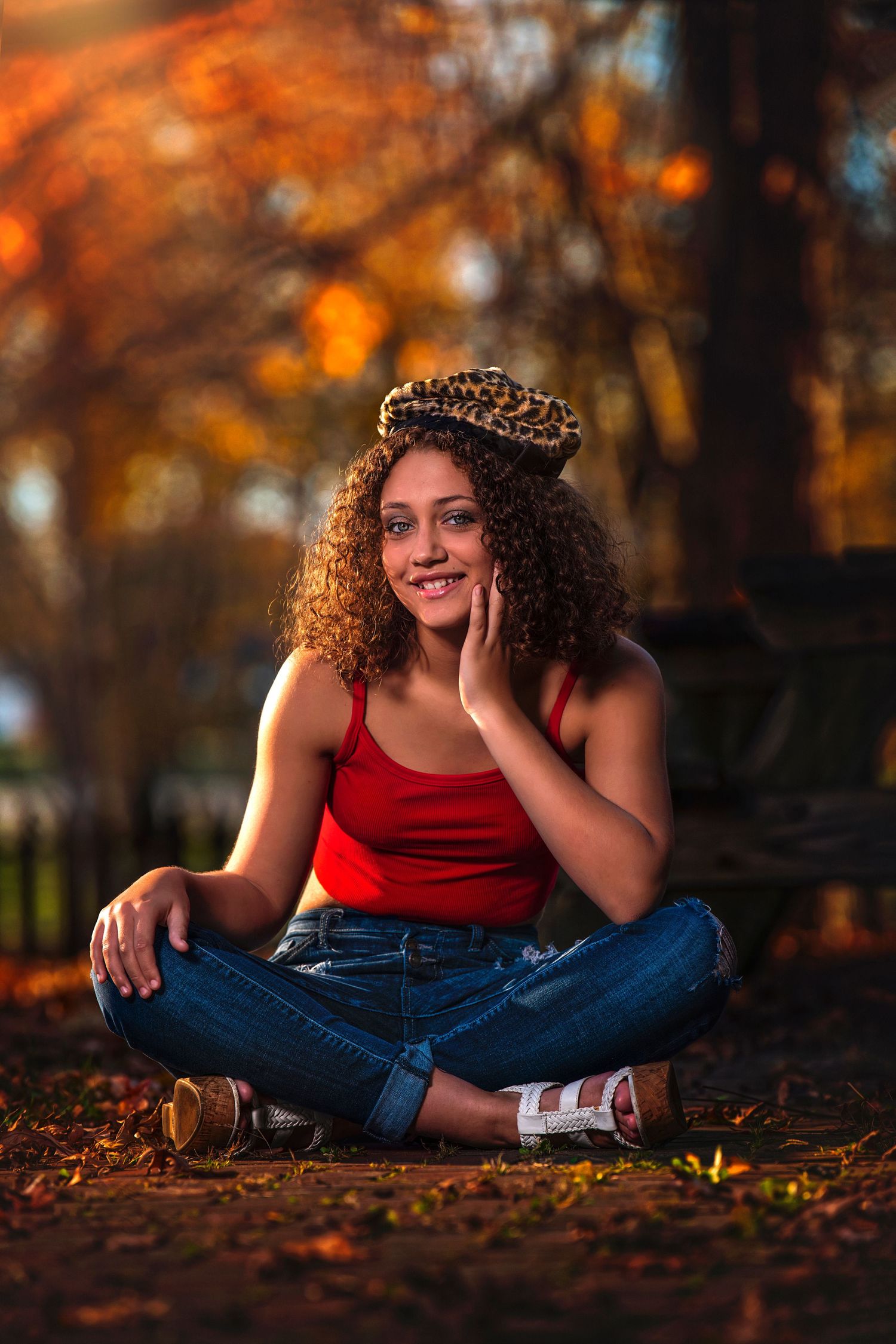 Young woman with curly hair and leopard beret sitting cross-legged in a park during golden autumn foliage.