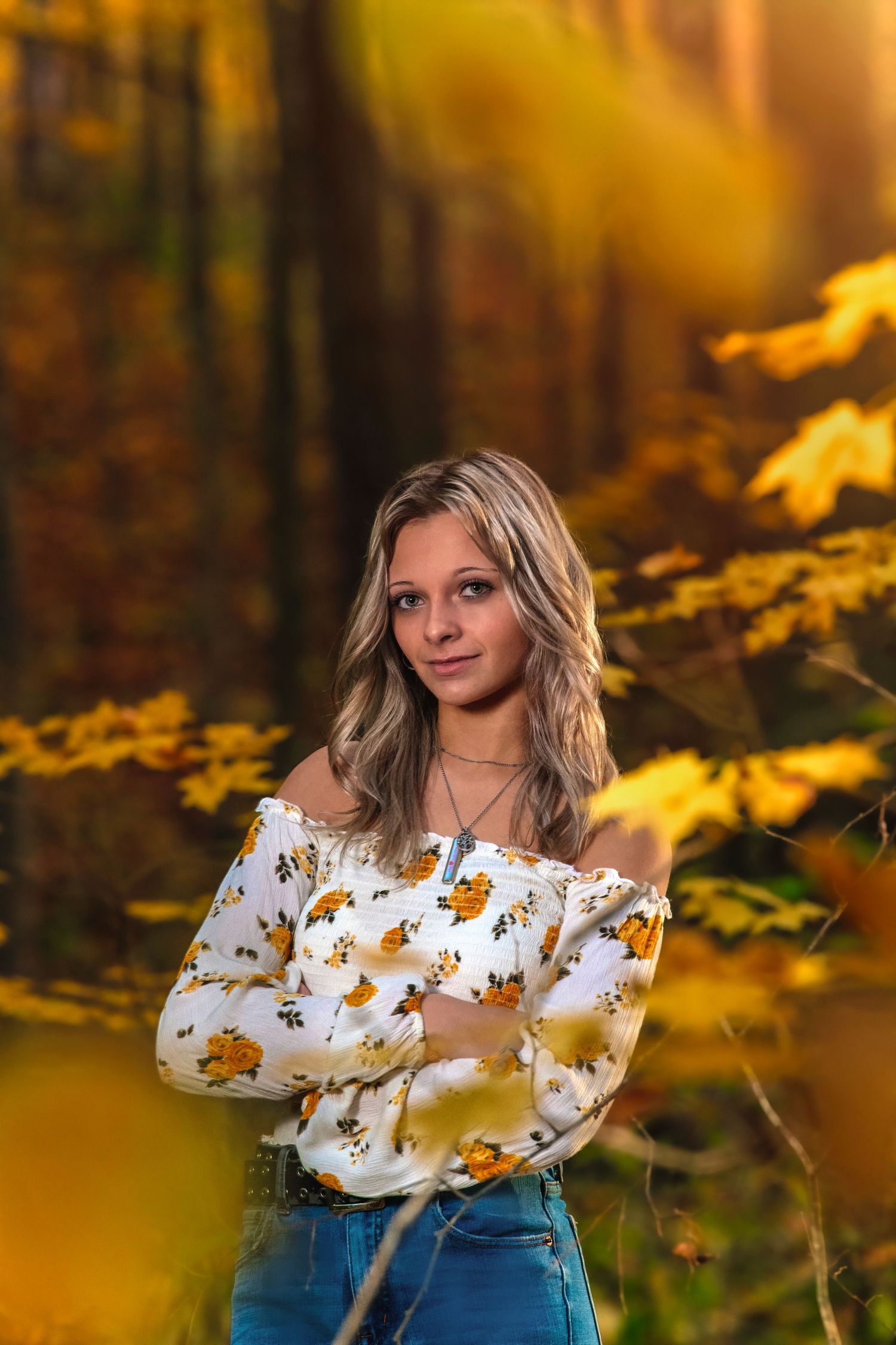 Young blonde woman in floral off-shoulder top posing in autumn forest with golden leaves.