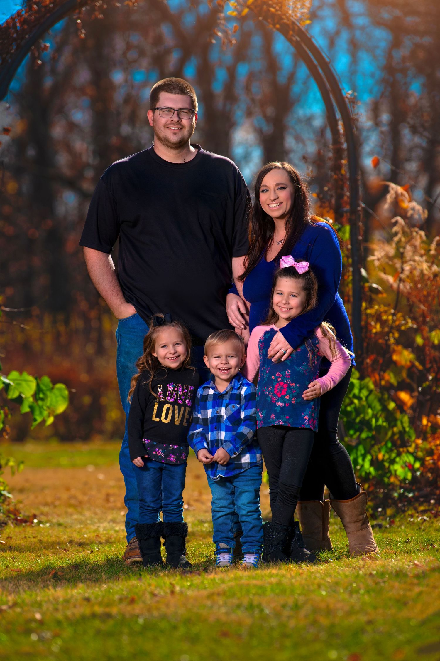Family of five posing outdoors in autumn setting with decorative arch, parents and three young children smiling.