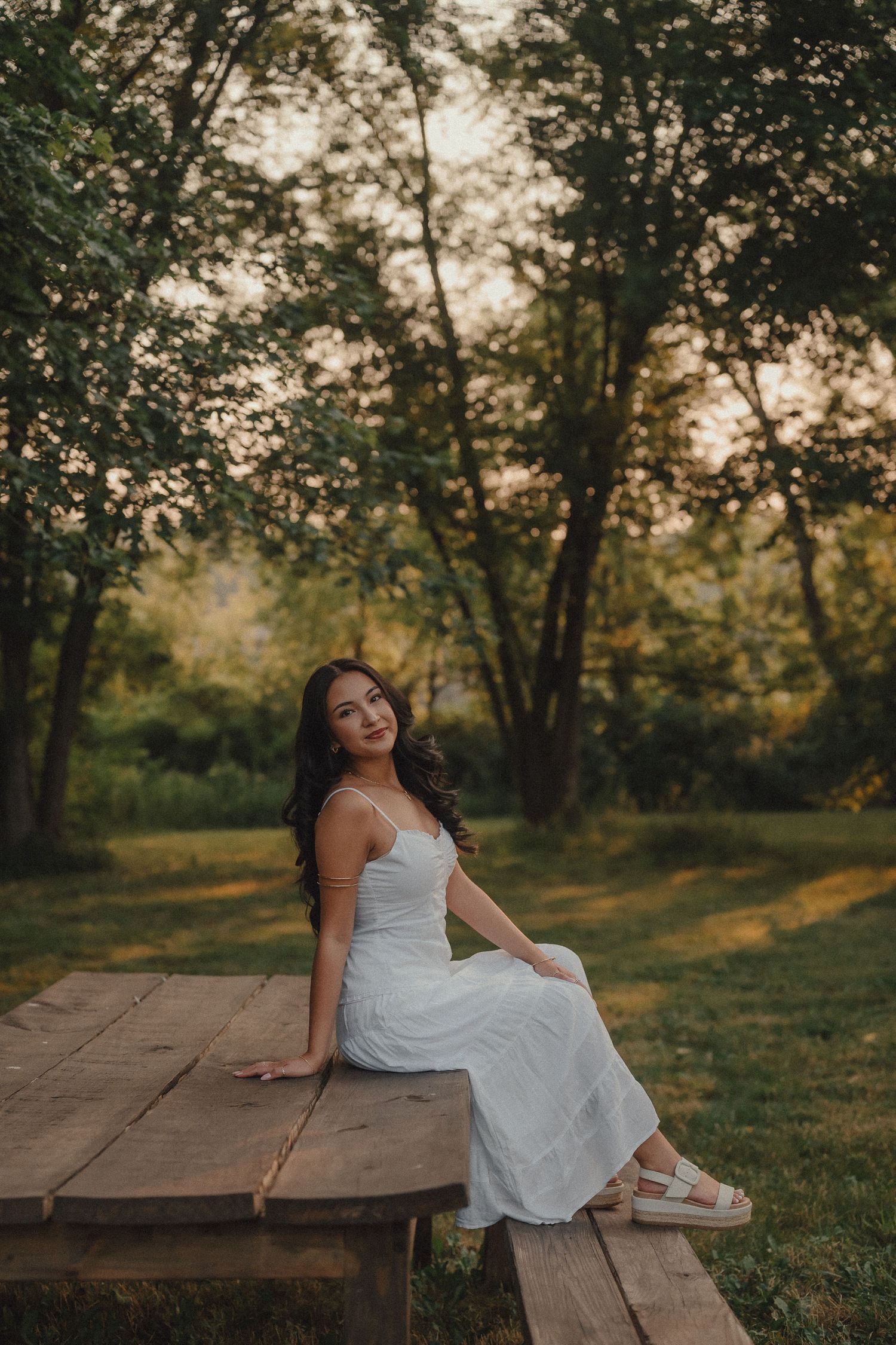 Young woman in white dress sitting on wooden table in lush green park at sunset.