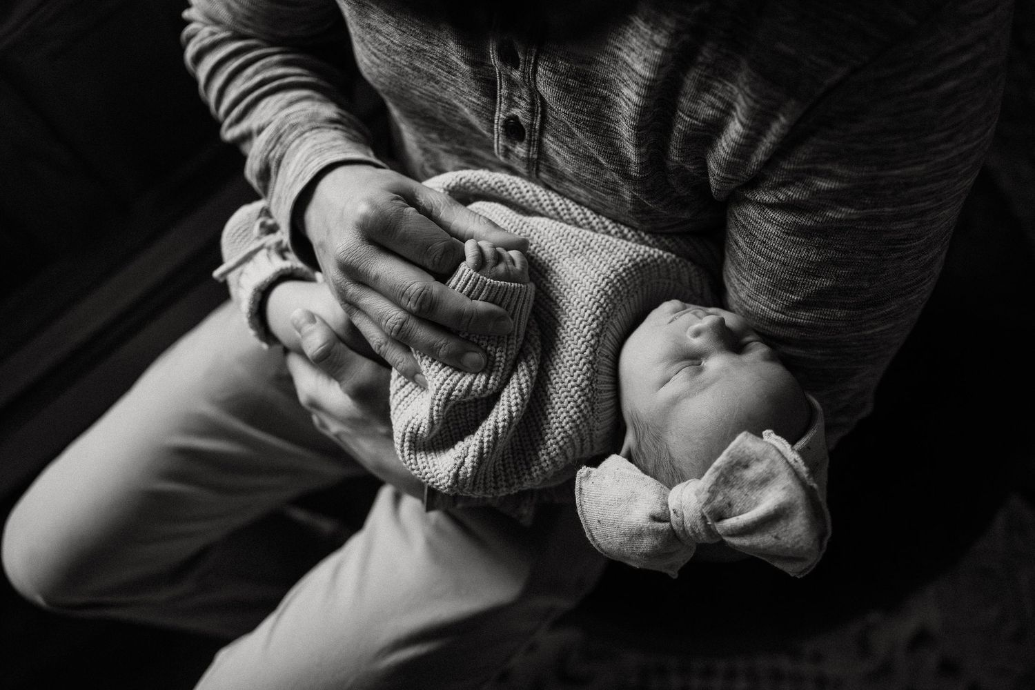 Black and white close-up of hands tenderly holding a swaddled infant against a dark background.