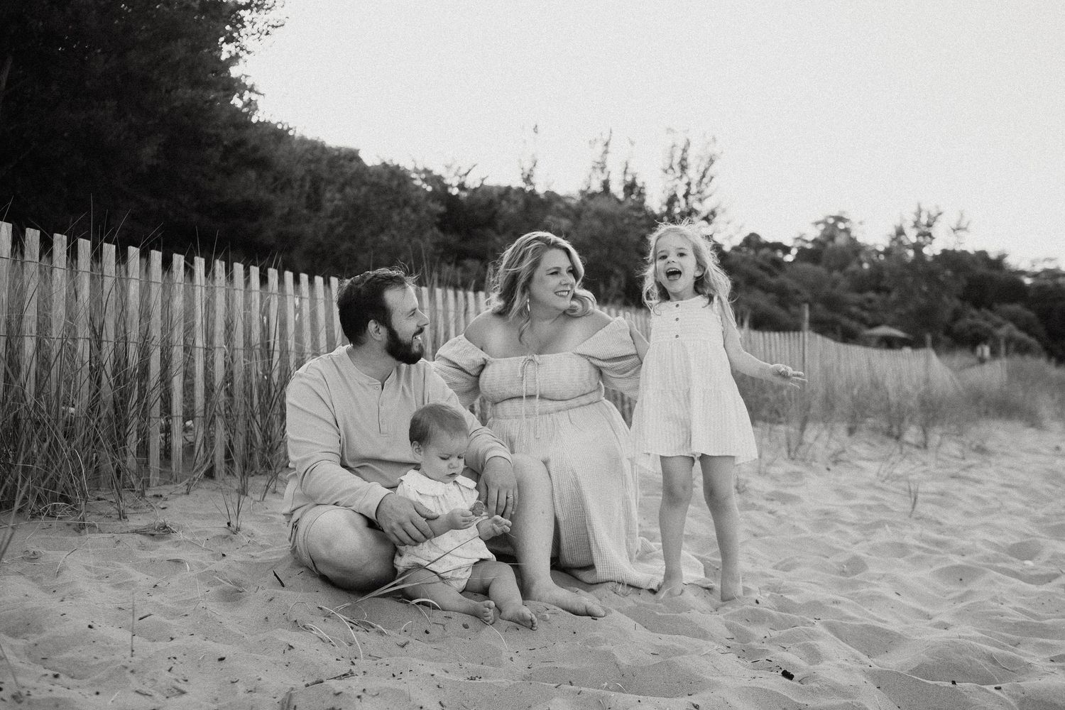 Black and white photo of family of four sitting on beach sand near wooden fence.