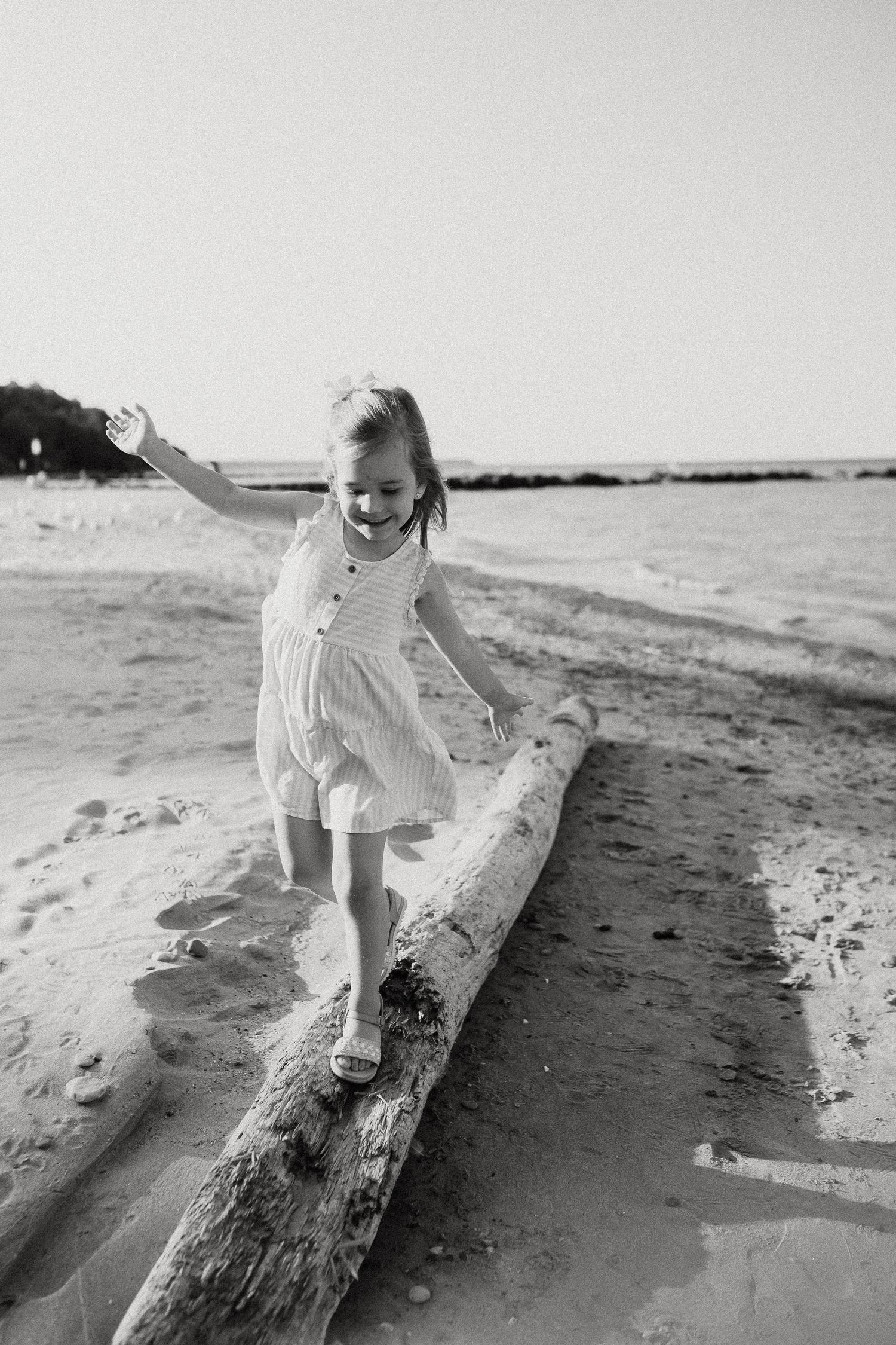 A young girl balances on a driftwood log on a sandy beach in a black and white photo.