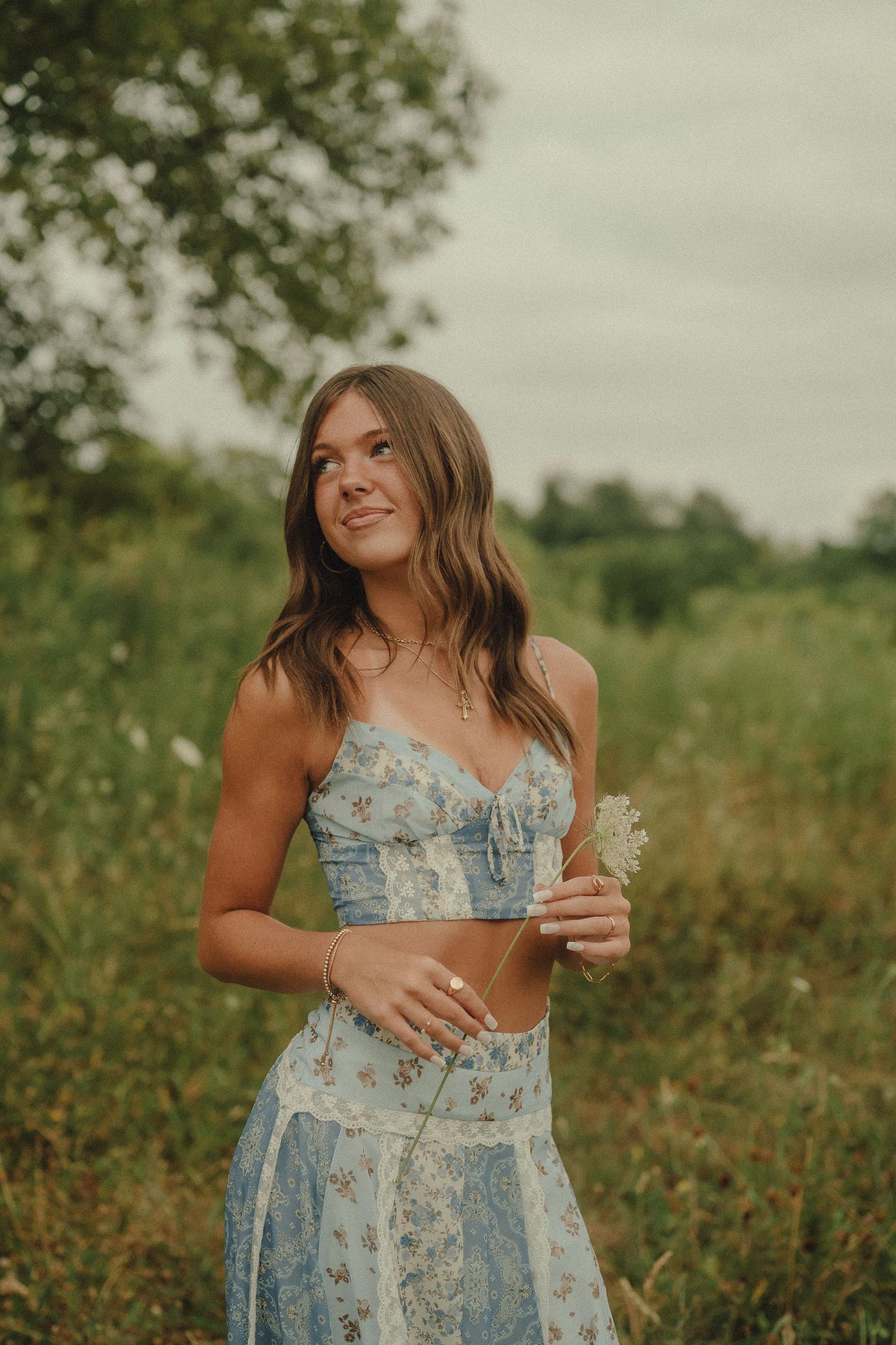 Young woman in blue floral two-piece set holding a wildflower while standing in a green meadow.