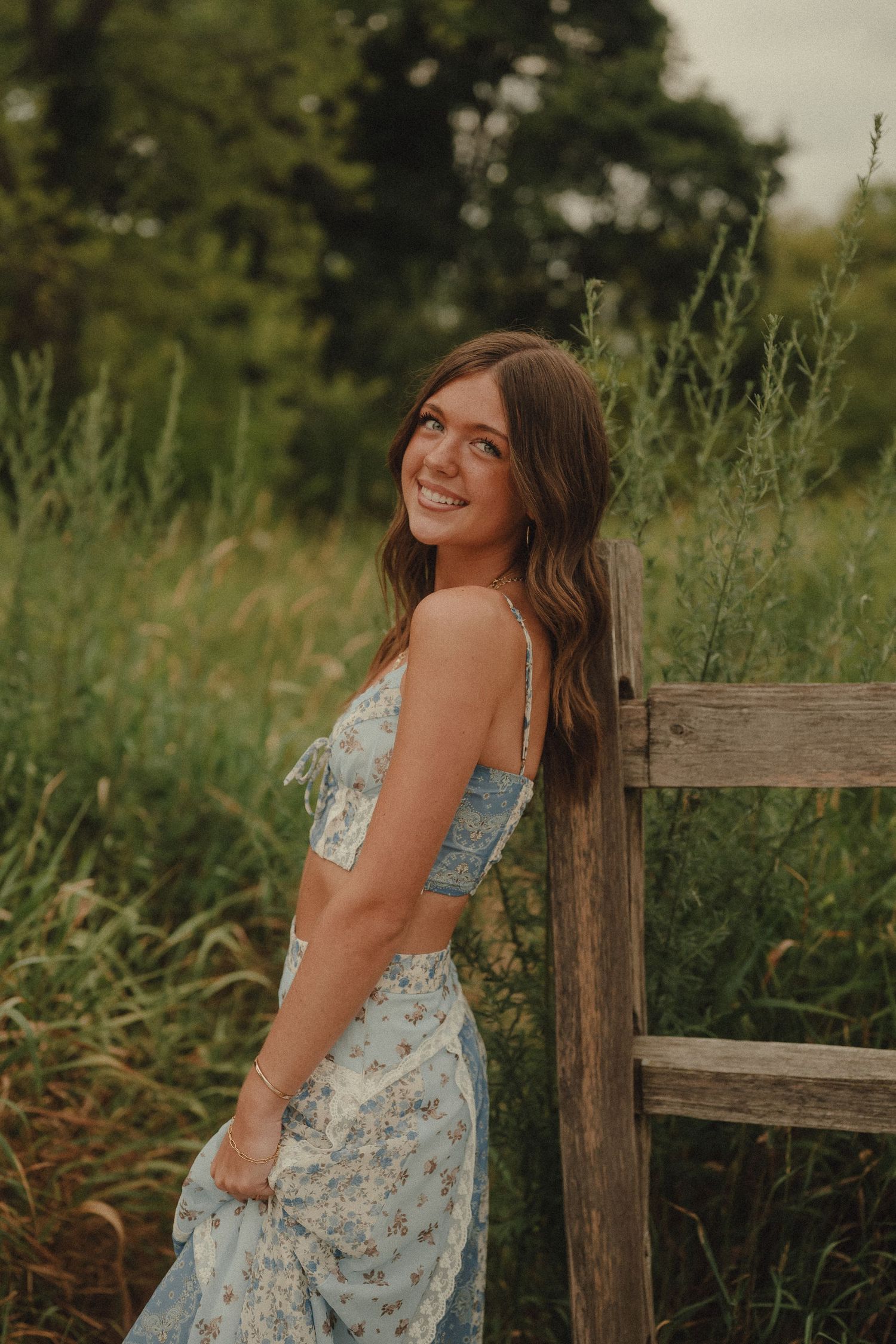 Young woman in blue floral dress smiling against rustic wooden fence in lush green field.