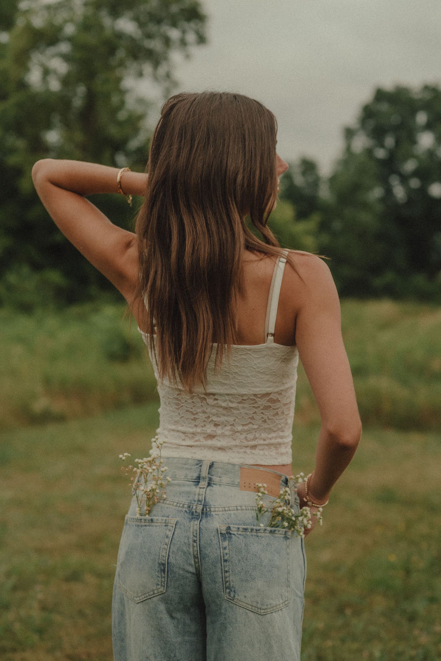 Back view of young woman in white lace top and jeans holding wildflowers in green field.