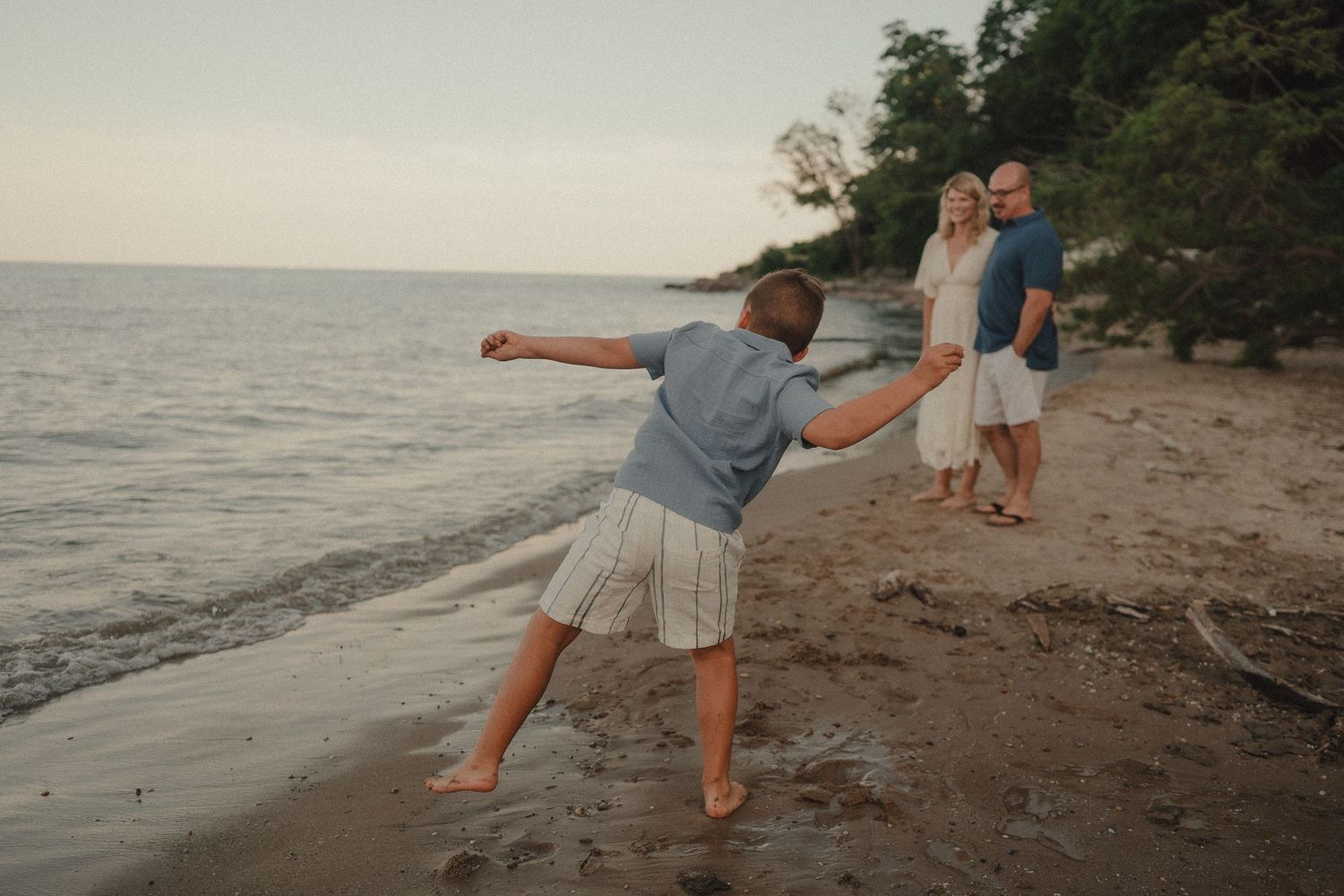 Child throwing a stone into the ocean while parents watch on a sandy beach at dusk.