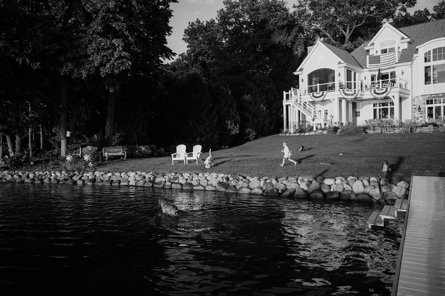 Children play on a rocky shoreline in front of a large lakeside home in black and white.
