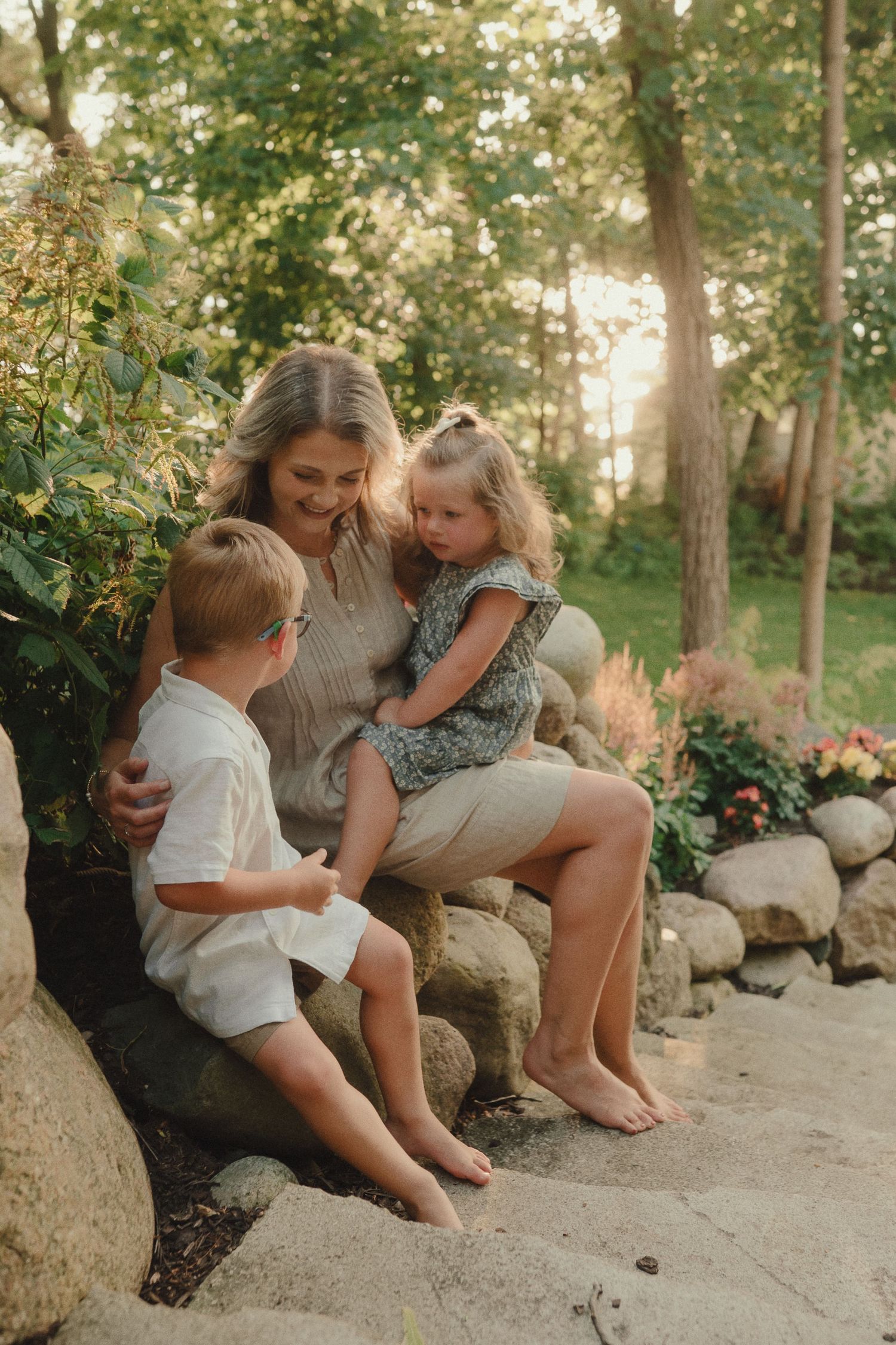 Mother sitting on stone wall with two young children in a sunlit garden, sharing a tender family moment.